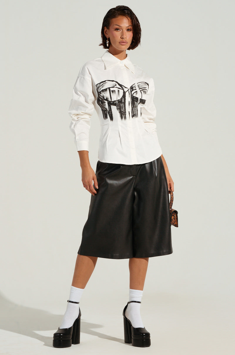 A person stands confidently in the BAD FOR YOU BUTTON DOWN BLOUSE, featuring a bustier-inspired graphic. They pair it with black leather culottes and black platform shoes, accentuated by white socks. A small handbag complements their look, along with short, styled hair against a plain background.