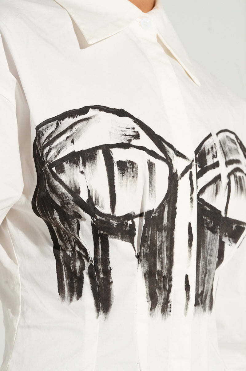 A close-up of the BAD FOR YOU BUTTON DOWN BLOUSE highlights a painted black abstract design that resembles a bustier-inspired graphic on the front. This white blouse is made from stretchy cotton and includes a classic collar for added style.