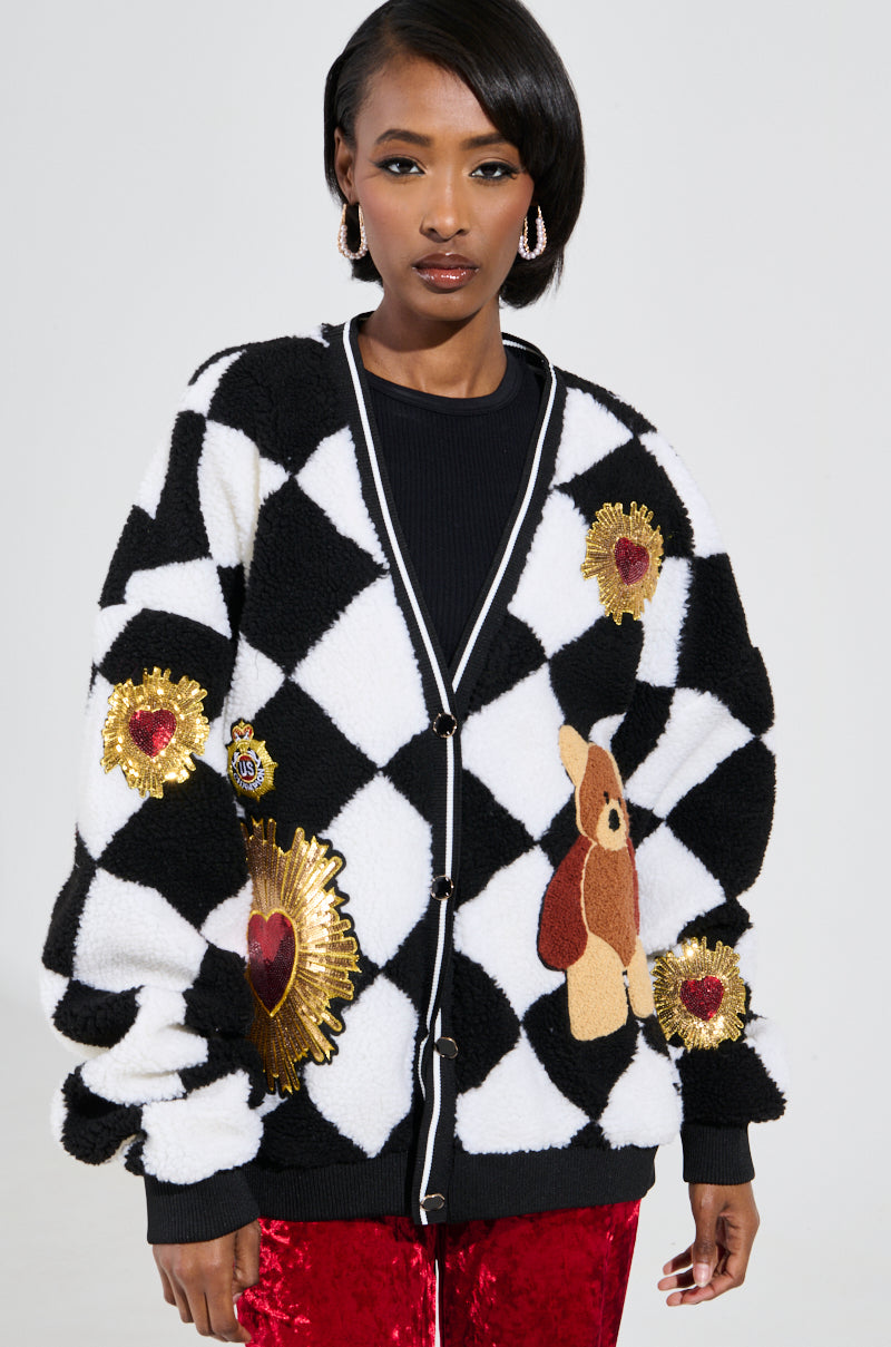 Dressed in the adorable TEDDY BEAR CUTE CHECKERED BUTTON DOWN CARDIGAN adorned with faux sherpa heart and teddy bear graphics, a person with short hair and hoop earrings pairs it perfectly with red velvet pants. The simple gray backdrop enhances the charm of their stylish outfit.