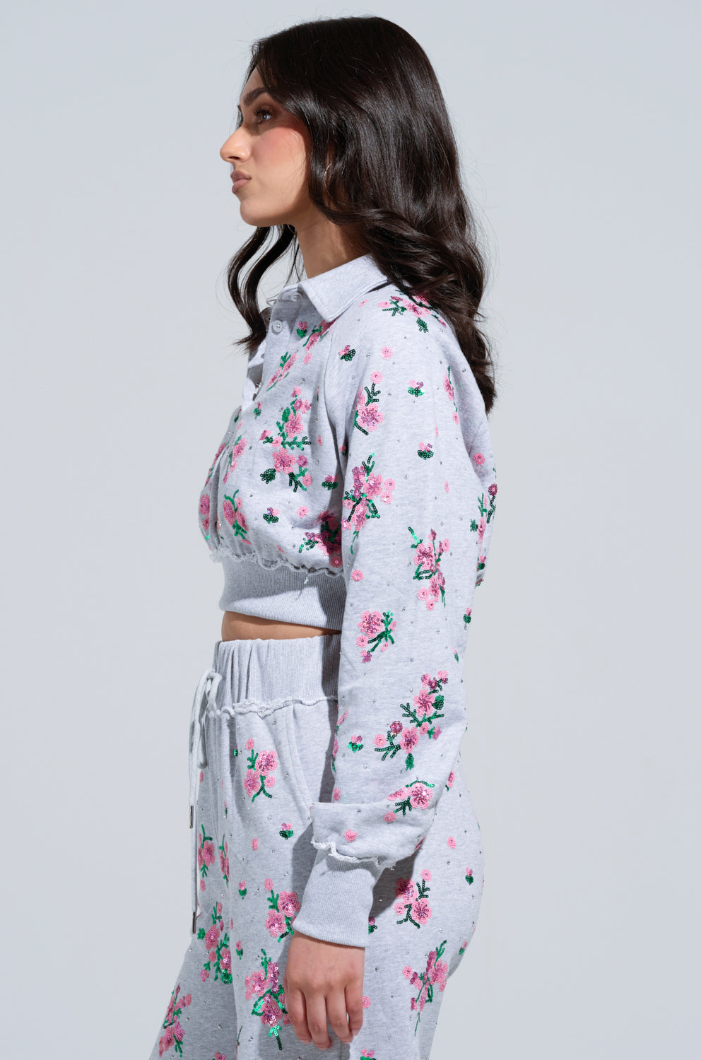 A person with long dark hair is wearing a grey matching set adorned with floral appliques featuring pink and green flowers. The outfit includes the GARDEN PARTY RHINESTONE BUTTON UP TOP and high-waisted pants. They are standing sideways against a plain background.