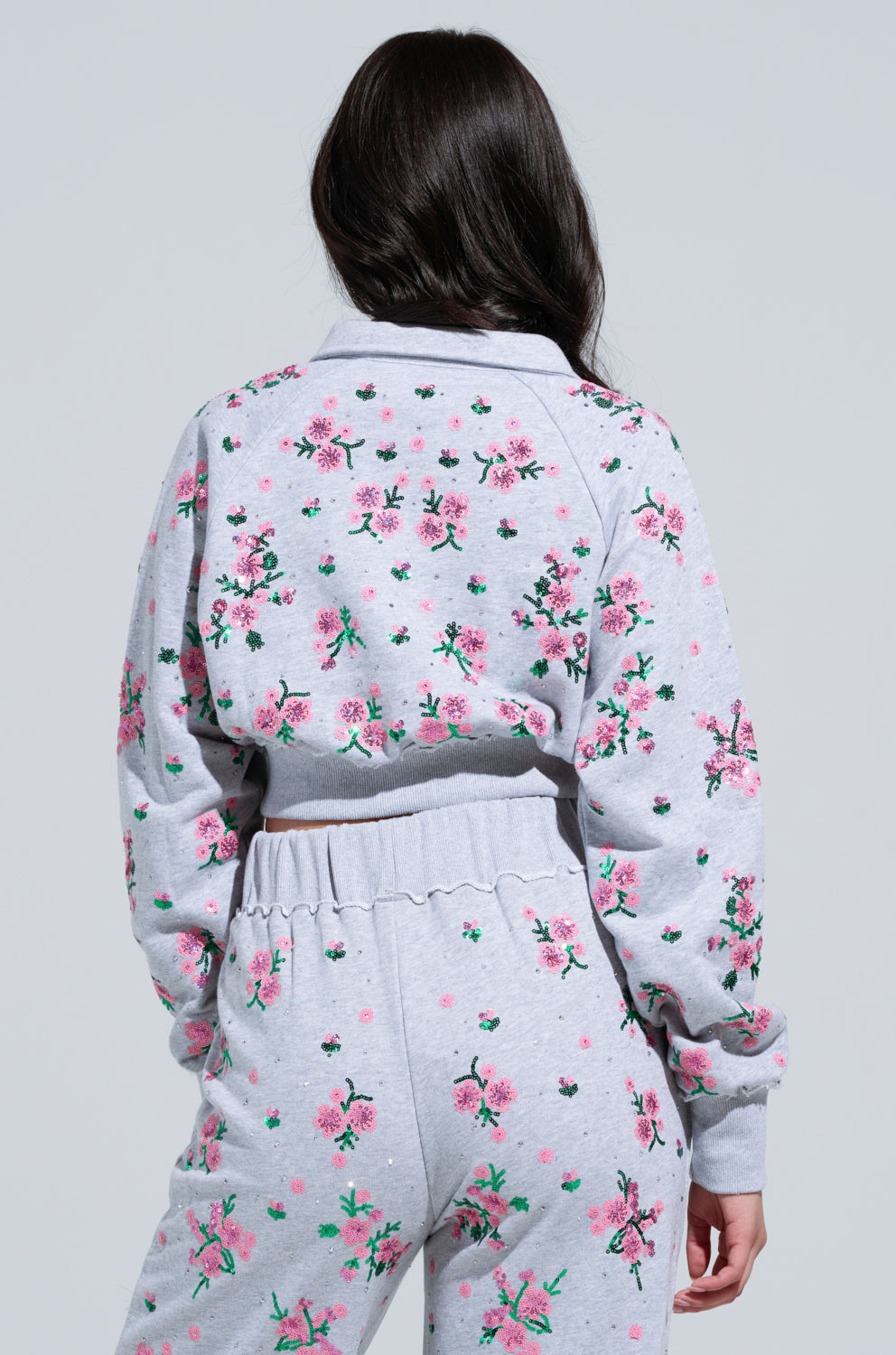 A person with long dark hair is wearing a matching gray Garden Party Rhinestone Button Up Top and pants set, adorned with pink and green floral appliques. The image shows their back as they stand against a light gray background.