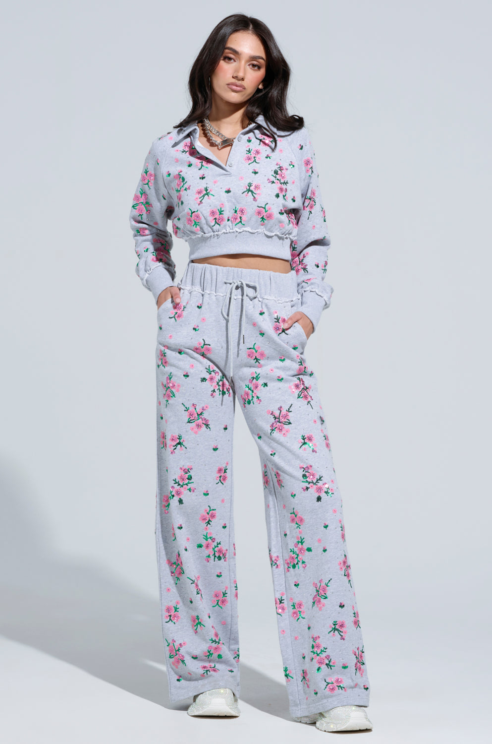 Dressed in the GARDEN PARTY RHINESTONE BUTTON UP TOP, featuring dazzling gray floral patterns and sparkling rhinestone buttons, a person poses confidently. The ensemble includes a cropped long-sleeve shirt paired with matching wide-leg pants adorned with pink and green flowers, complemented perfectly by white sneakers for a chic look.