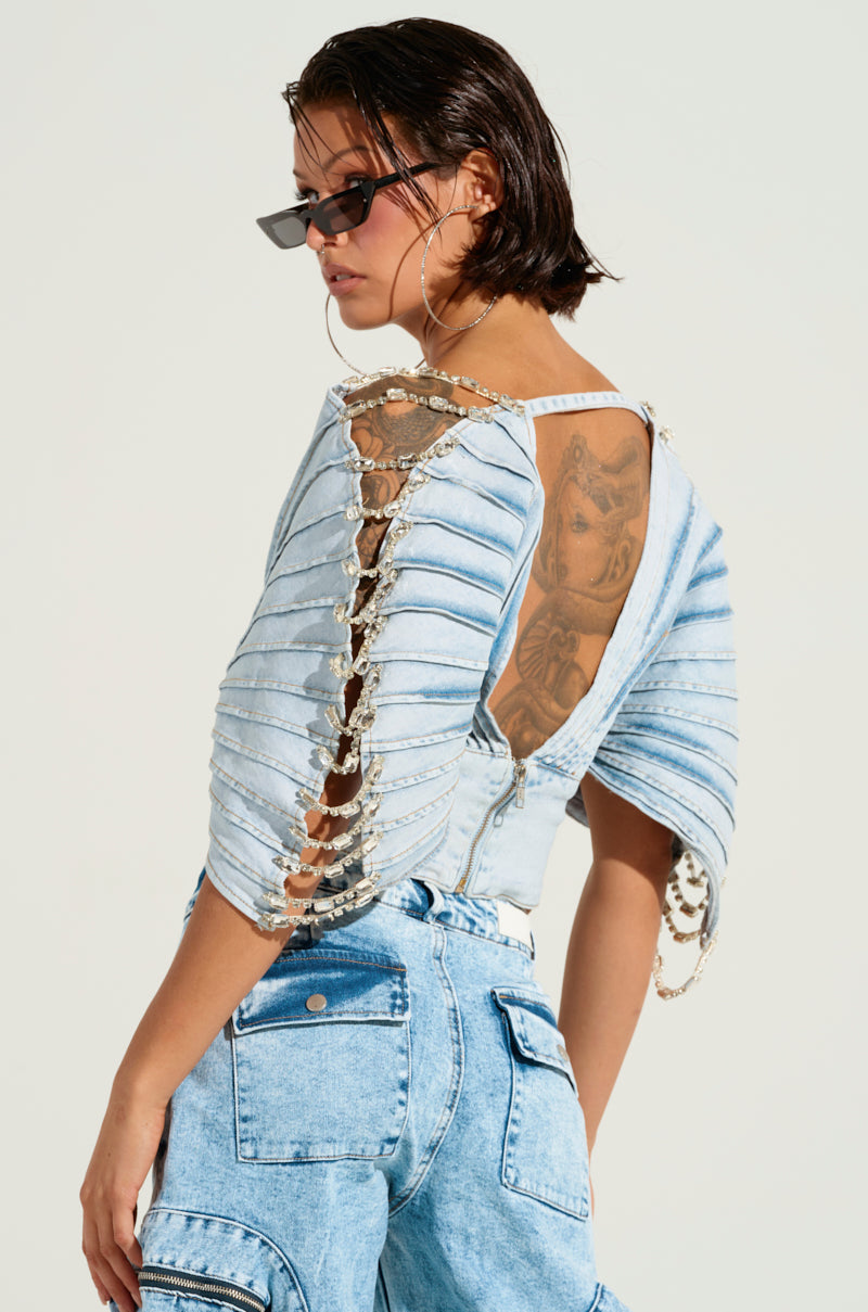 A person with short dark hair and sunglasses confidently poses in the ASHLEE RHINESTONE ENCRUSTED DENIM TOP, an off-the-shoulder piece featuring rhinestones and chain details. They complete the look with matching jeans that have pockets, revealing a tattoo on their arm.
