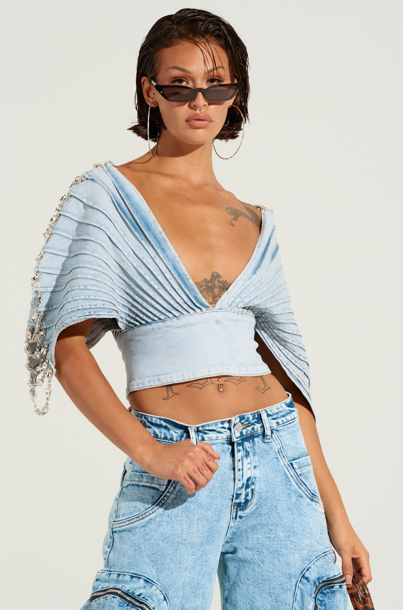 A person with short hair wears cat-eye sunglasses and an ASHLEE RHINESTONE ENCRUSTED DENIM TOP featuring pleated sleeves, paired with high-waisted denim pants. Rhinestone-encrusted tattoos shimmer on their chest and waist, set against a plain, light background.