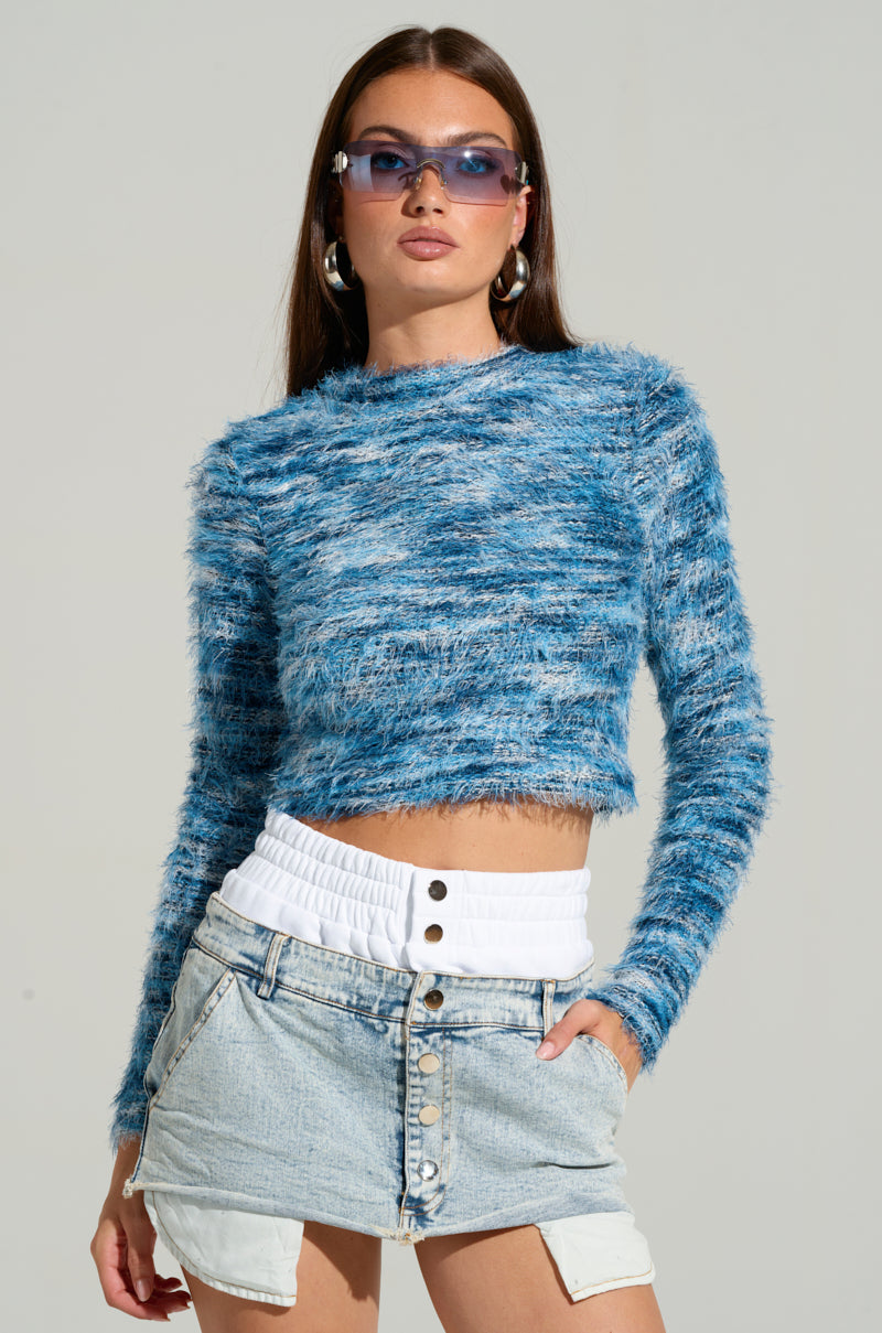 A person confidently poses against a plain background, wearing blue-tinted sunglasses and the BIG FUZZY ENERGY CROPPED SWEATER IN BLUE. They're dressed in a high-waisted blue denim skirt, have their hands in their pockets, and accessorize with large hoop earrings.