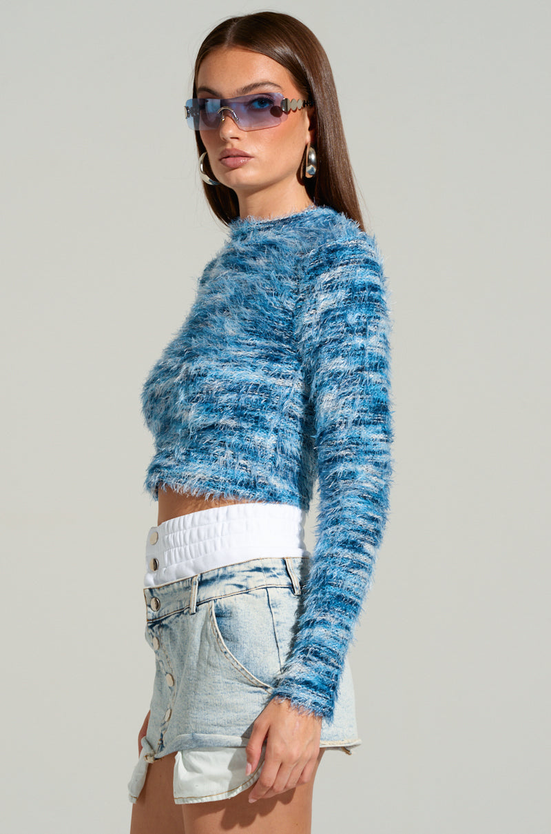Modeling a bold look, an individual wearing sunglasses and hoop earrings shows off the BIG FUZZY ENERGY CROPPED SWEATER IN BLUE. This mohair-inspired sweater, showcasing a wavy pattern design, is paired with light denim shorts featuring a white waistband and prominent buttons, all set against a simple light background.