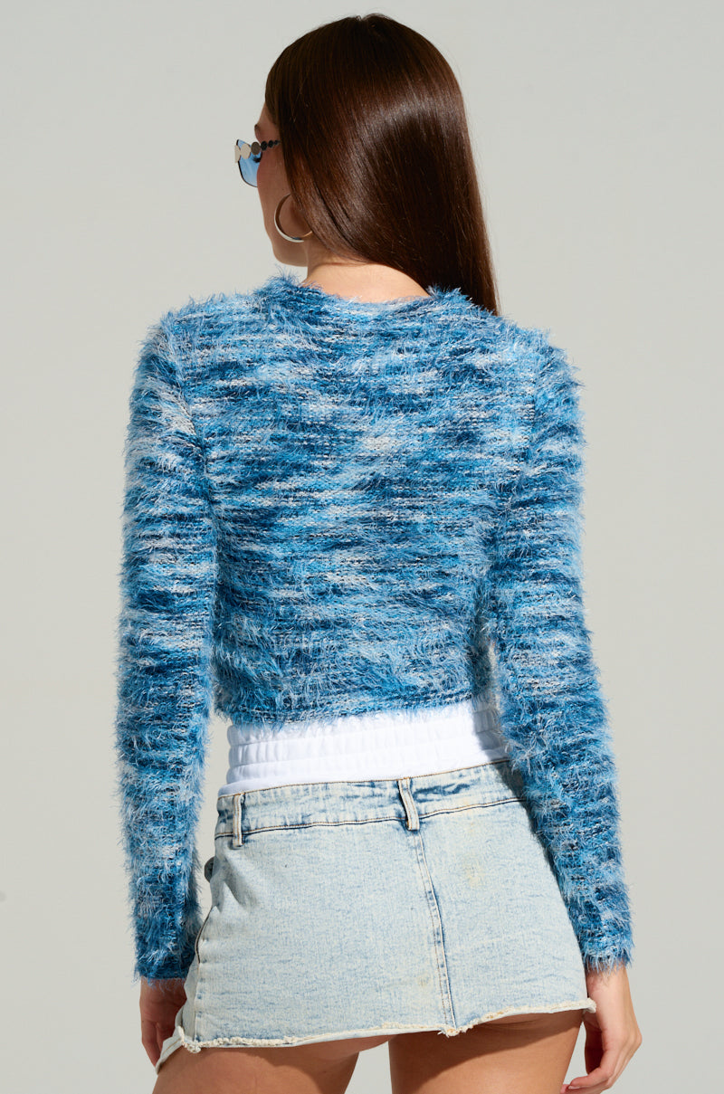 A person with long hair is wearing sunglasses, the BIG FUZZY ENERGY CROPPED SWEATER IN BLUE, and a short denim skirt. They are standing against a plain gray background, facing away from the camera.