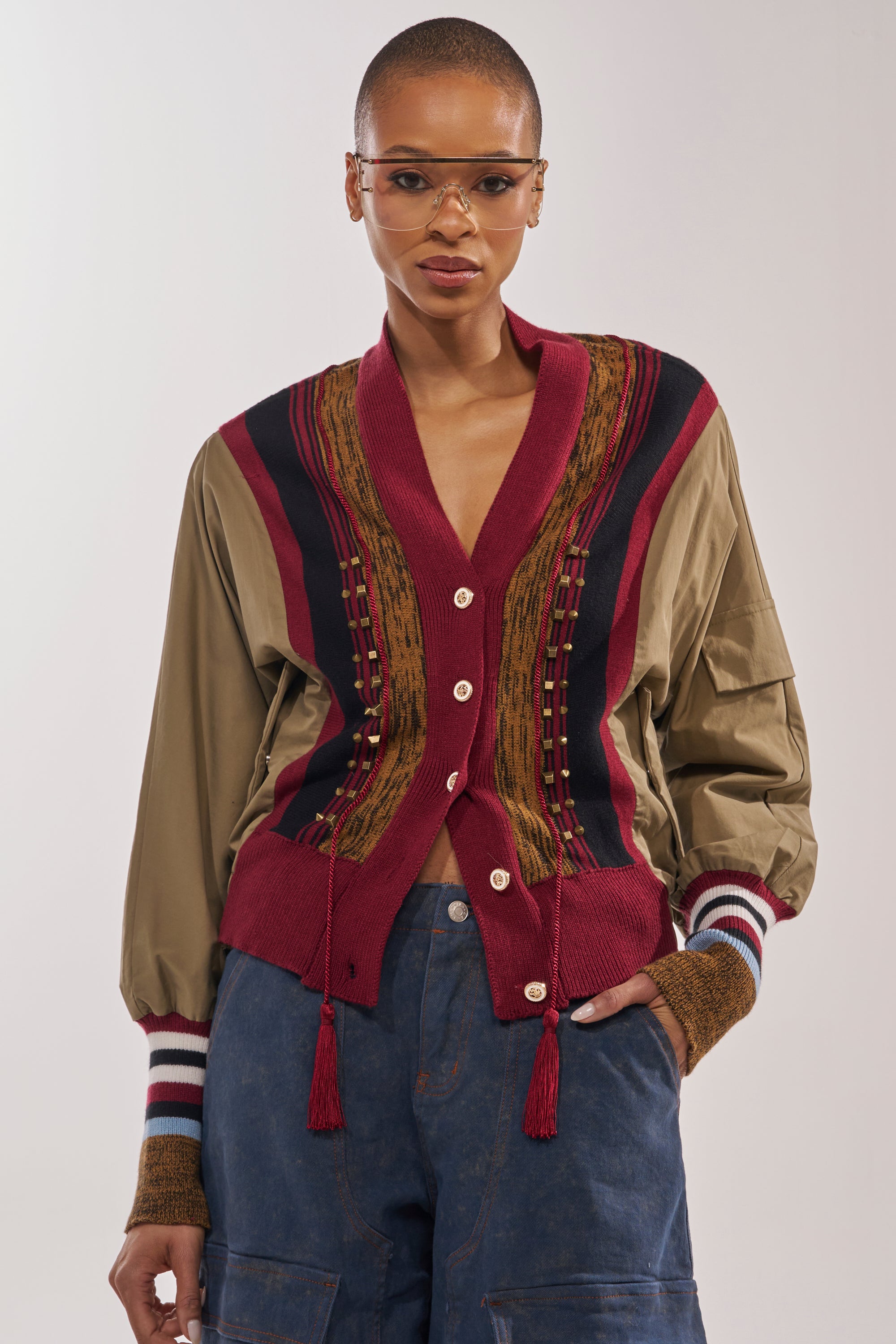 A person with short hair and clear glasses wears the NEW DAY COLORBLOCK CARDIGAN, featuring red, navy, and tan patterns, over a brown shirt and blue pants. They stand confidently against a plain background. 0