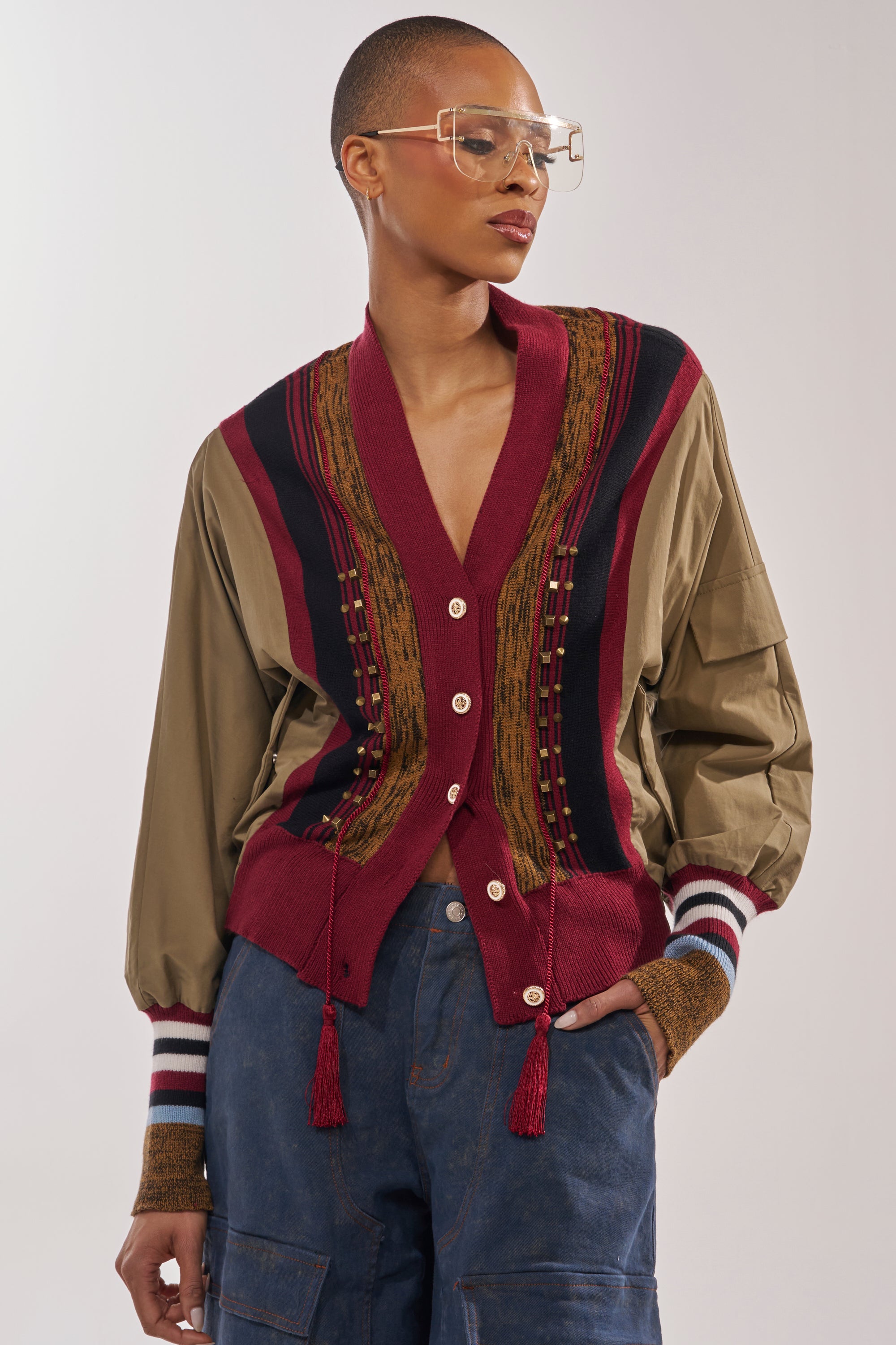A stylish person stands against a neutral background wearing clear glasses, the NEW DAY COLORBLOCK CARDIGAN with maroon and brown patterns and tassels over a tan shirt, and blue cargo pants, with one hand in their pocket. 1