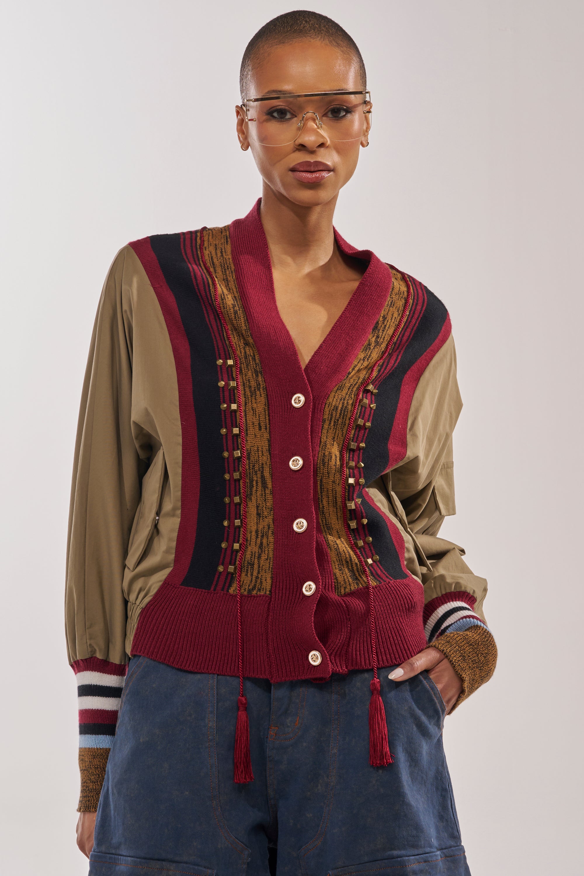 A person with short hair wears tinted glasses, loose blue pants, and the NEW DAY COLORBLOCK CARDIGAN in khaki and burgundy with buttons and textured details. One hand is in their pocket as they stand against a neutral background. 2