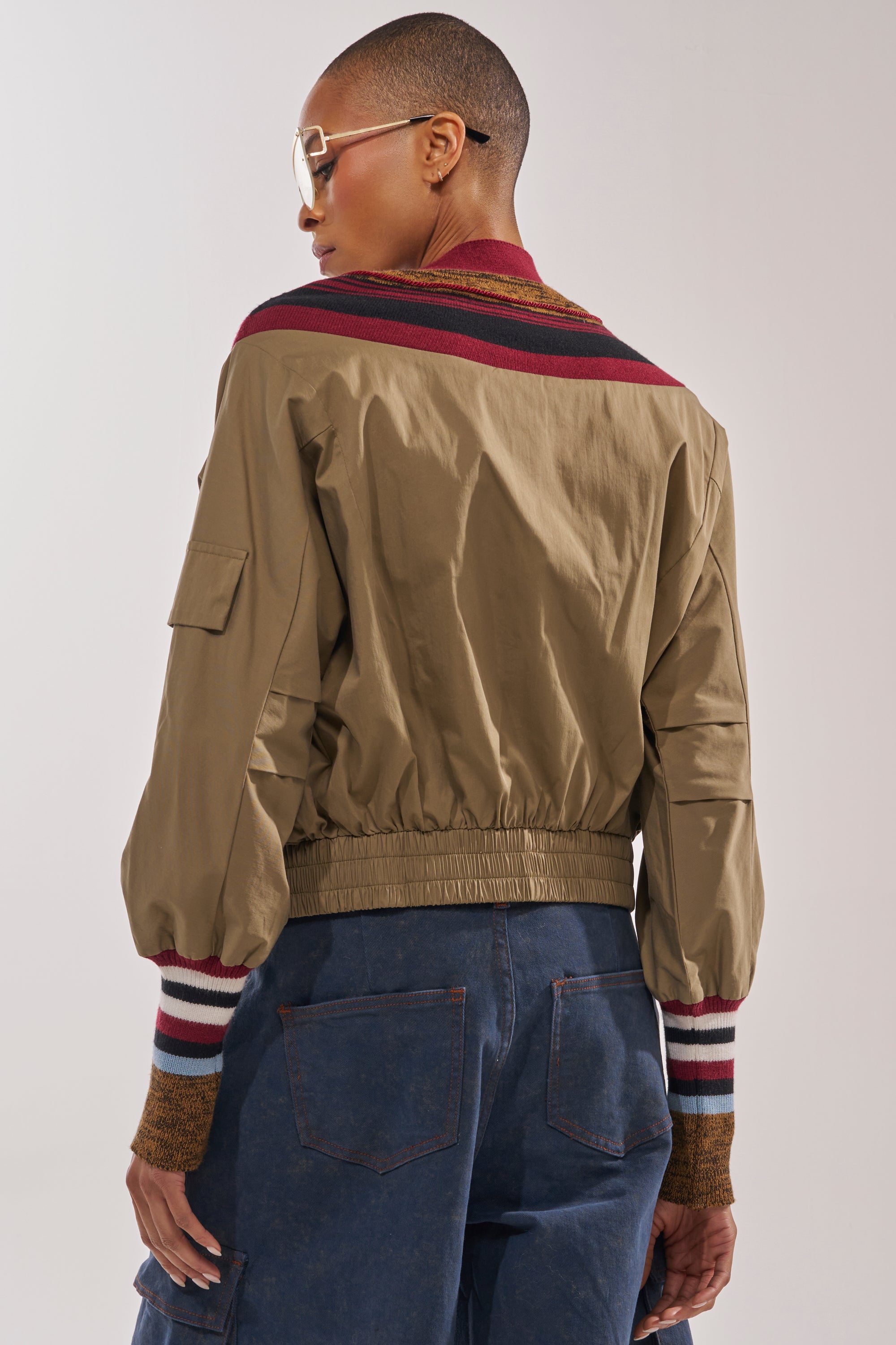 A person with short hair and glasses, wearing the NEW DAY COLORBLOCK CARDIGAN—a khaki bomber-style piece with maroon and white striped trim—and dark blue jeans, stands facing away from the camera. 4