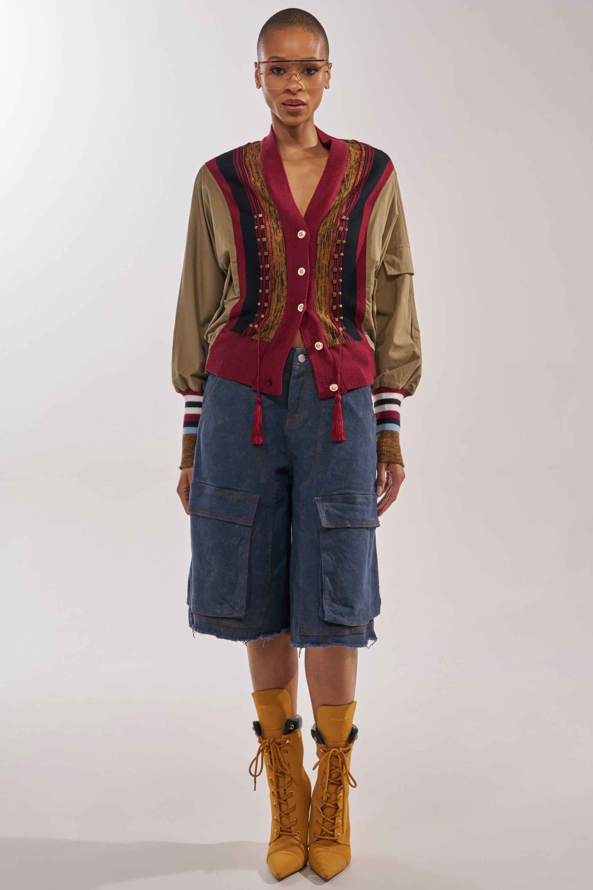 A person stands facing forward in the NEW DAY COLORBLOCK CARDIGAN with red and tan buttons and tassels, blue cargo knee-length shorts, yellow lace-up boots, and clear glasses against a plain light background. 5