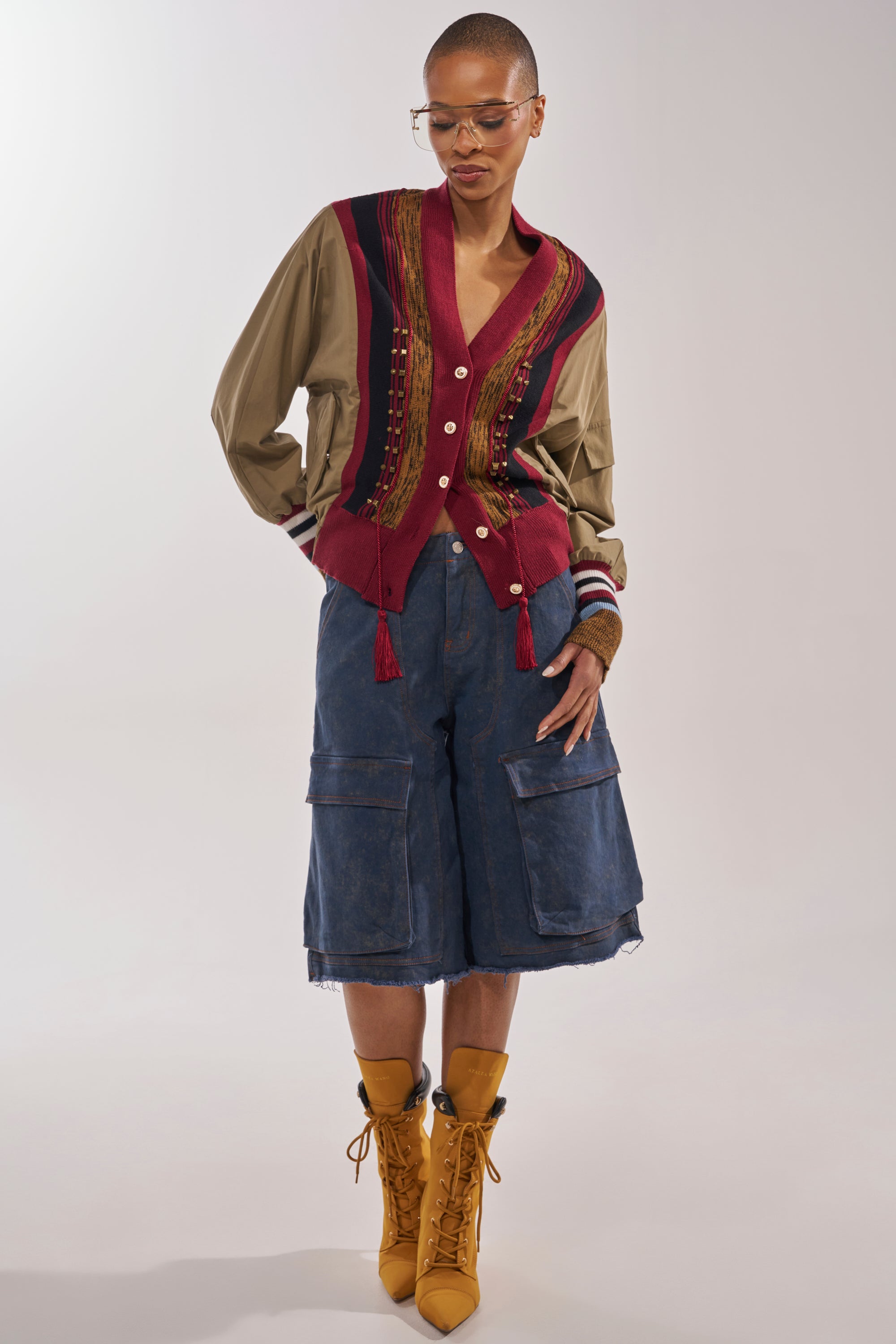 A person with short hair wears clear glasses, the NEW DAY COLORBLOCK CARDIGAN in red and tan, loose denim cargo shorts, and tan lace-up boots, standing hands-in-pockets against a neutral background. 6