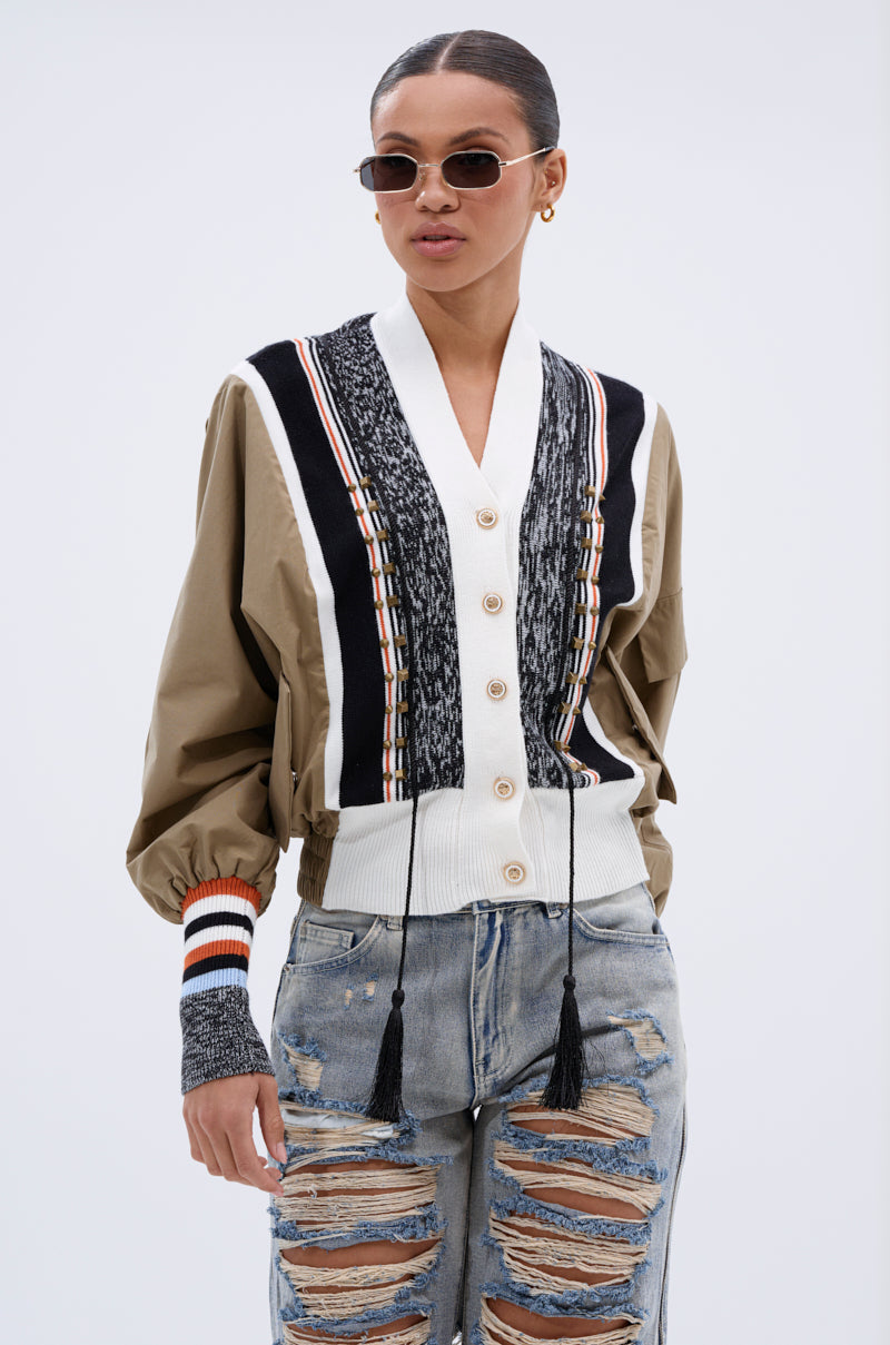 A woman stands against a plain light background wearing sunglasses, distressed light blue jeans, and the BEAUTIFUL DAY CARDIGAN—a color-blocked sweater with a white collar and tassels.