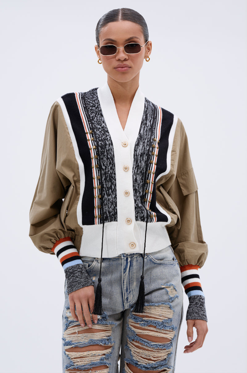 A woman wearing the BEAUTIFUL DAY CARDIGAN—a color-blocked cardigan—over a tan shirt with balloon sleeves, paired with distressed ripped jeans, stands against a plain light gray background. She's accessorized with round sunglasses and gold hoop earrings.