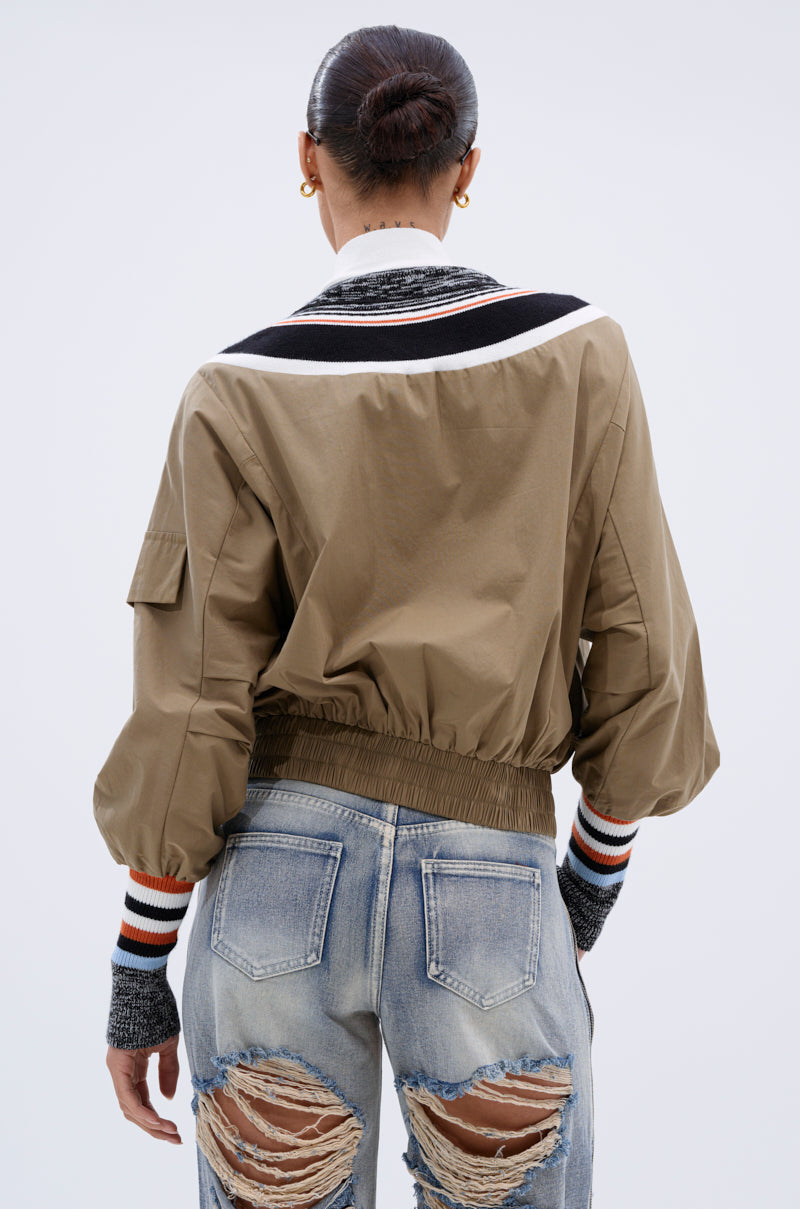 A person with hair in a bun wears the BEAUTIFUL DAY CARDIGAN, featuring tan color-blocking and striped cuffs and collar, paired with light blue distressed jeans. The photo is taken from behind against a plain background.