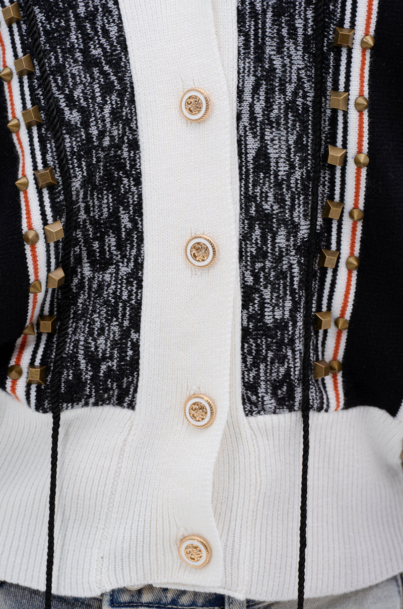 Close-up of the BEAUTIFUL DAY CARDIGAN, a color-blocked knit with black, white, and patterned panels, six beige buttons, gold square studs, red and white placket stripes. Blue denim jeans are also seen at the bottom.