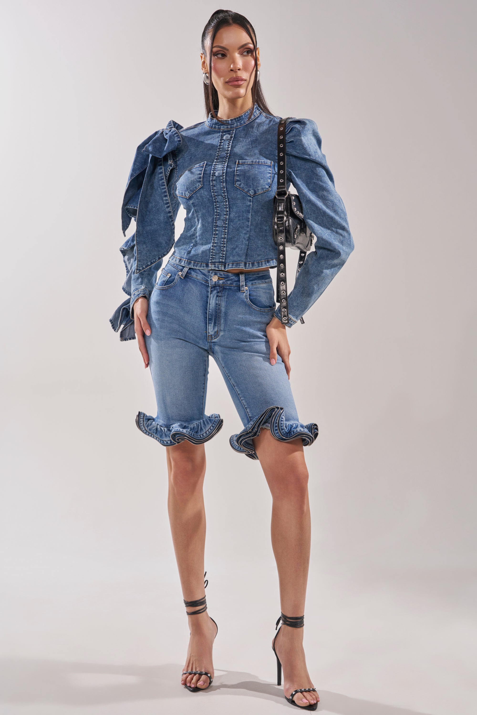 A woman stands in a studio wearing the SEIZE THE DAY LONG SLEEVE FLOUNCE MEDIUM BLUE DENIM TOP paired with matching knee-length denim shorts, holding a studded black handbag and wearing black high-heeled sandals.
