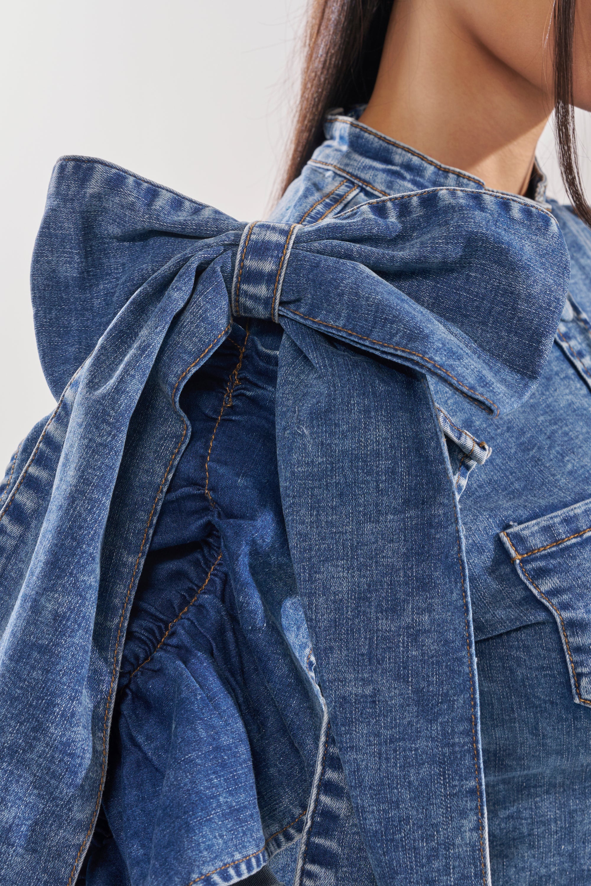 A close-up of a person wearing the SEIZE THE DAY LONG SLEEVE FLOUNCE MEDIUM BLUE DENIM TOP, featuring a matching large bow shoulder, visible stitching, and crafted from blue denim fabric.