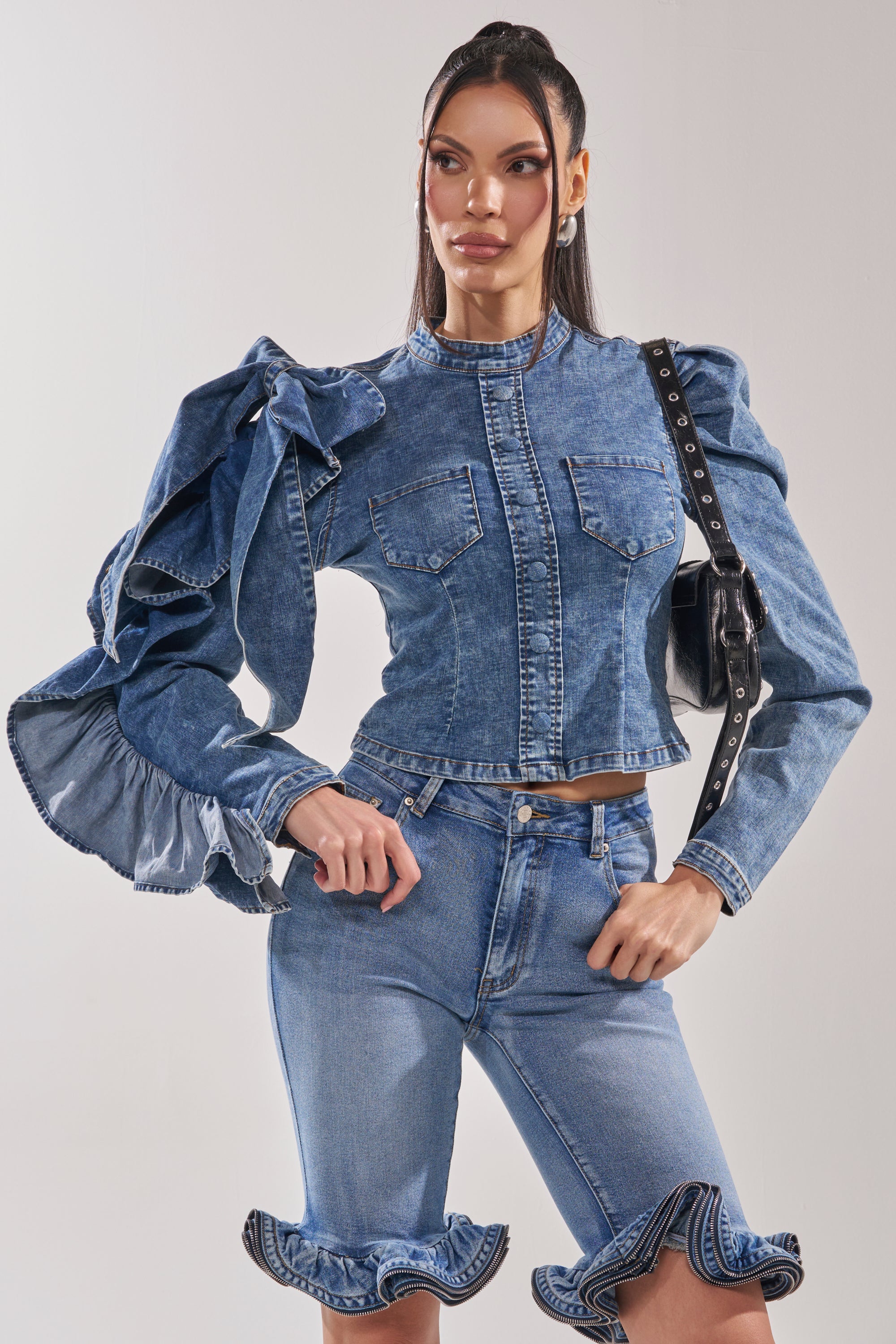 A woman confidently poses in the SEIZE THE DAY LONG SLEEVE FLOUNCE MEDIUM BLUE DENIM TOP, featuring dramatic ruffled sleeves, paired with matching high-waisted jeans and a black shoulder bag.
