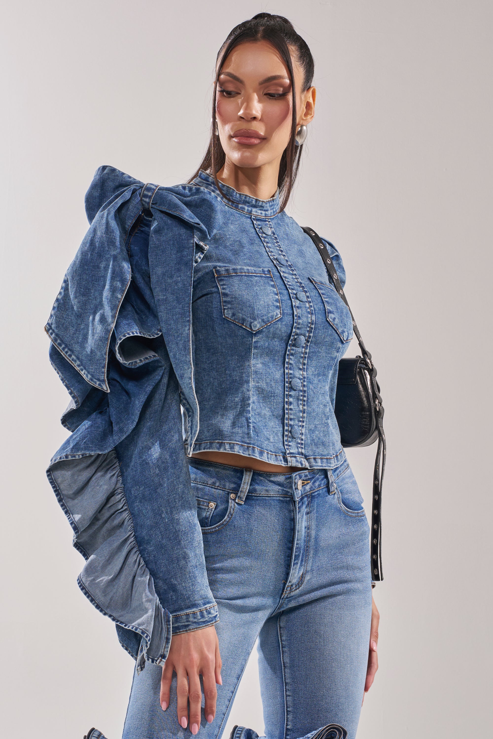 A woman with long dark hair and subtle makeup poses against a plain background wearing the SEIZE THE DAY LONG SLEEVE FLOUNCE MEDIUM BLUE DENIM TOP, styled with matching blue jeans and a black shoulder bag.