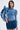 A woman wears the SEIZE THE DAY LONG SLEEVE FLOUNCE MEDIUM BLUE DENIM TOP with a mini skirt—an undeniably fashion-forward look featuring exaggerated ruffles at one shoulder. She has straight dark hair, hoop earrings, and stands against a plain light background.