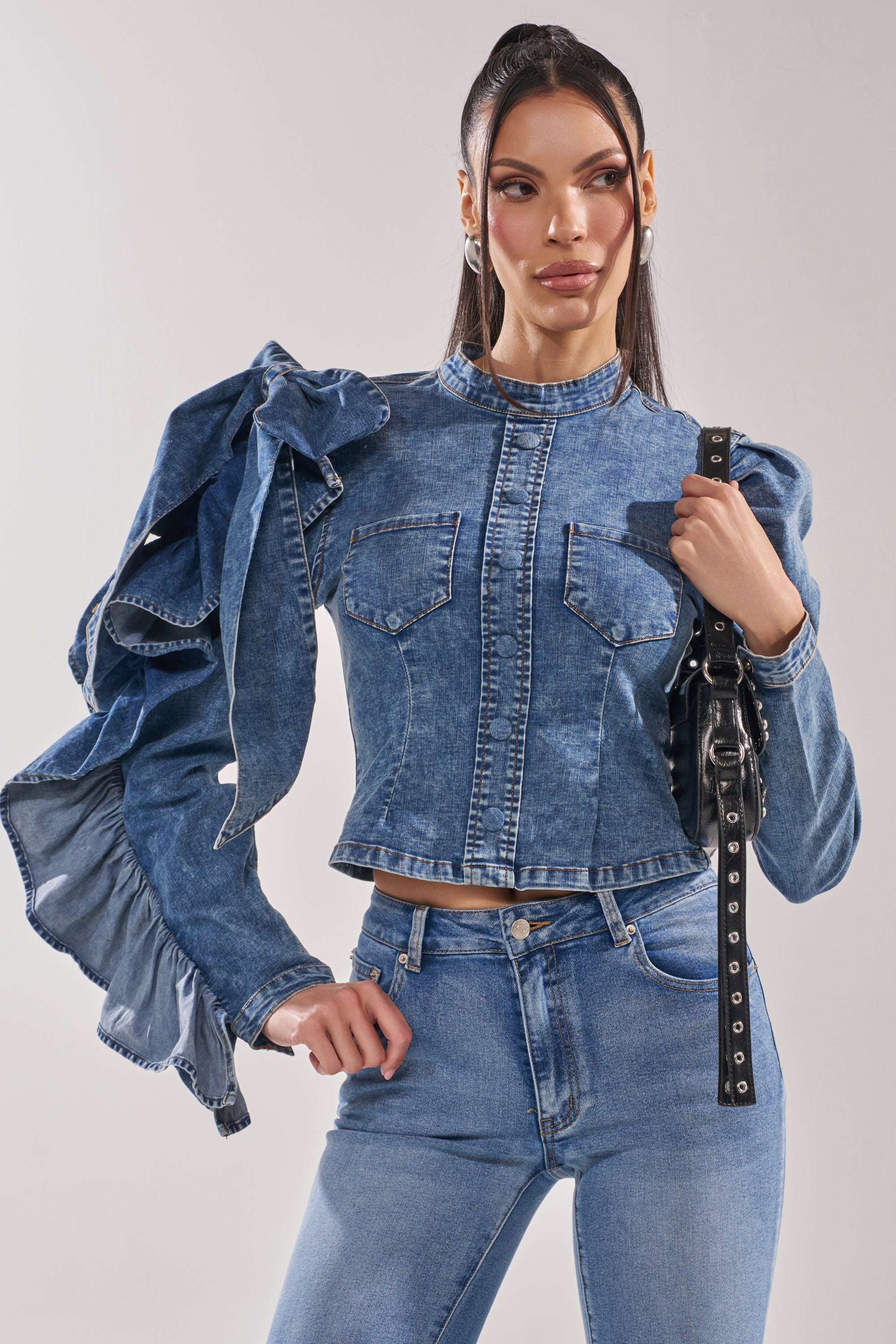 A woman poses confidently against a plain background, wearing the SEIZE THE DAY LONG SLEEVE FLOUNCE MEDIUM BLUE DENIM TOP with dramatic puffed sleeves and matching high-waisted jeans, accessorized with a black studded shoulder bag.
