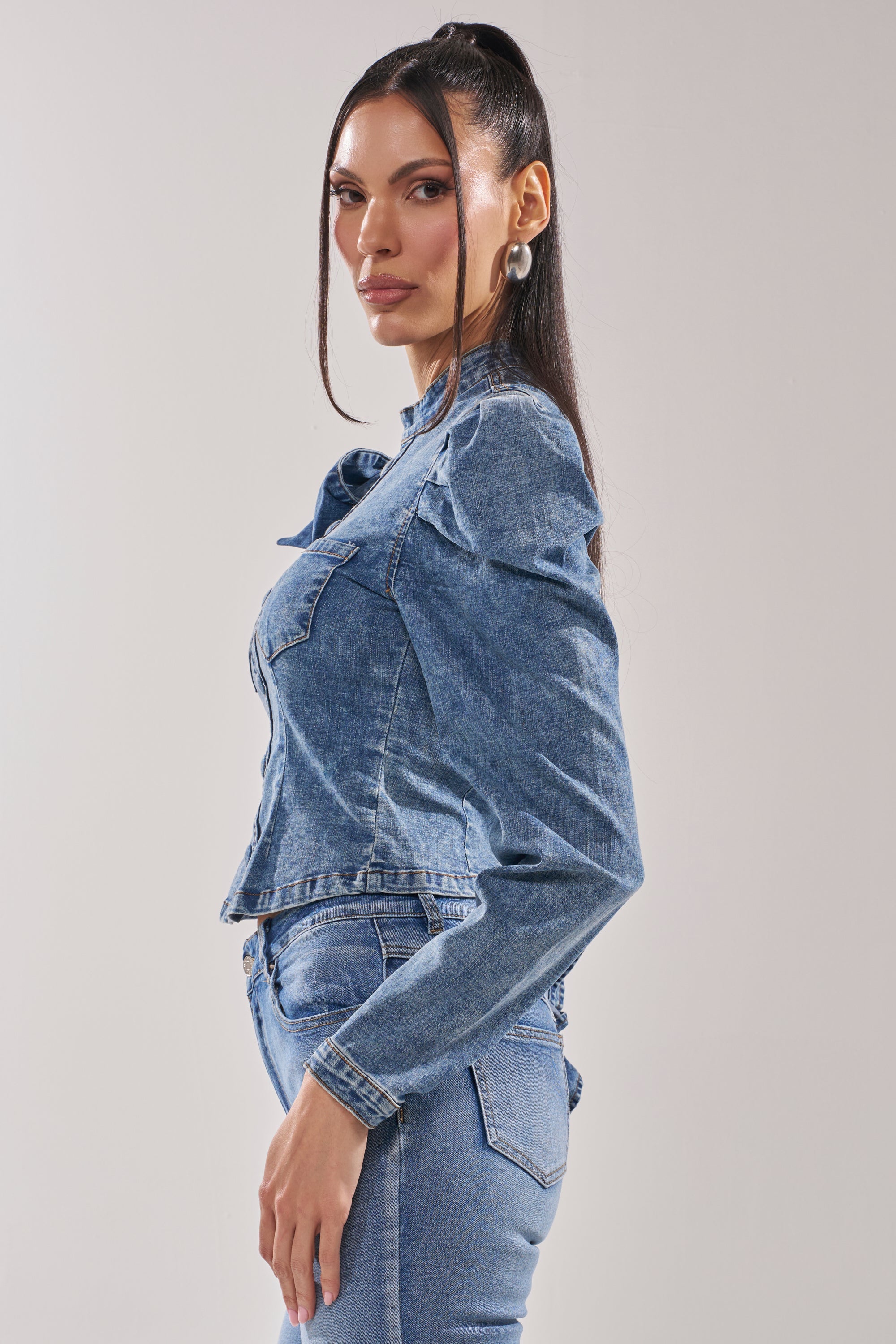 A woman with long dark hair stands sideways against a plain light background, wearing the SEIZE THE DAY LONG SLEEVE FLOUNCE MEDIUM BLUE DENIM TOP and jeans, looking at the camera with a neutral expression.