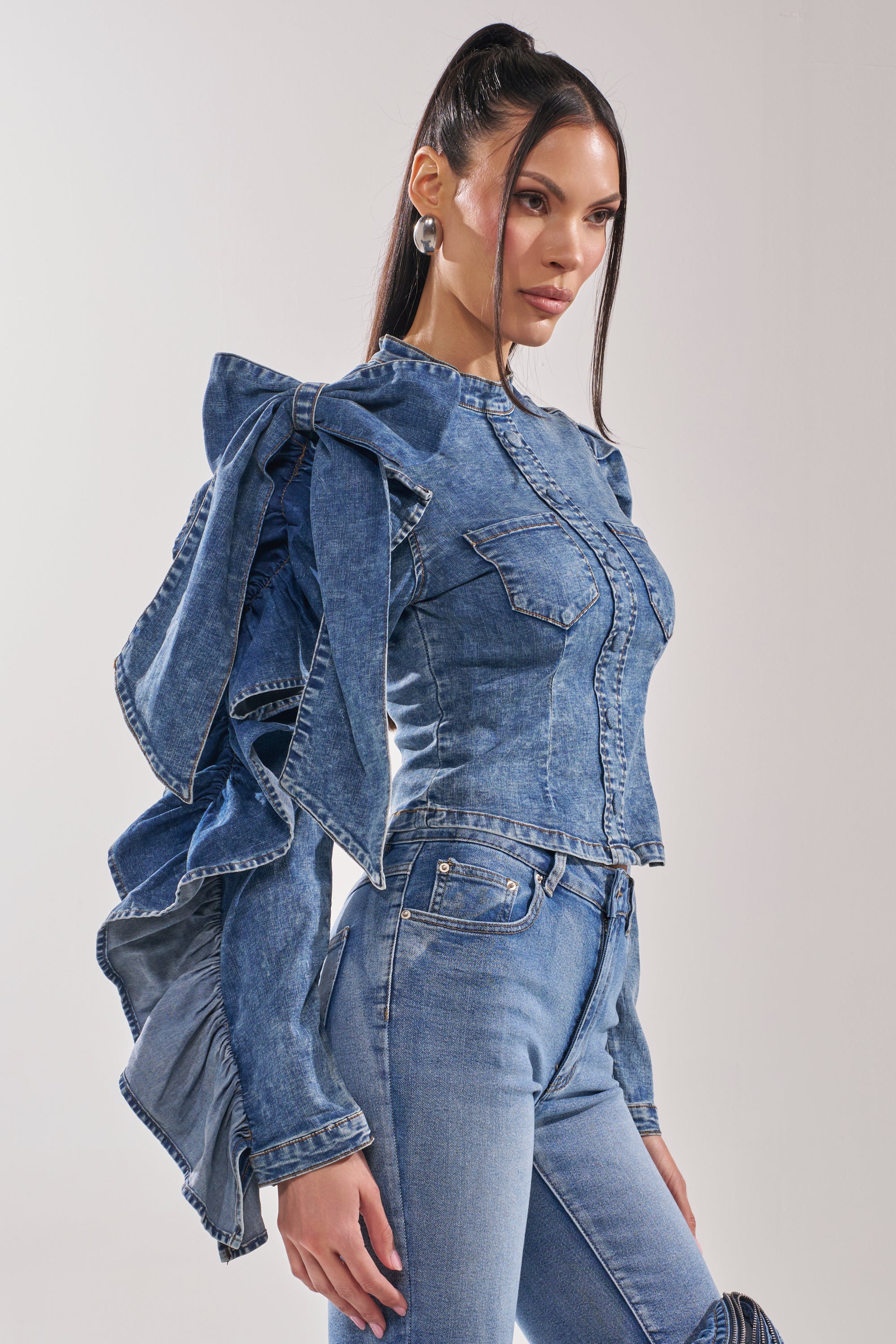 A woman wears the SEIZE THE DAY LONG SLEEVE FLOUNCE MEDIUM BLUE DENIM TOP with dramatic ruffled sleeves and matching light-wash jeans, standing against a plain background and looking confidently to the side.