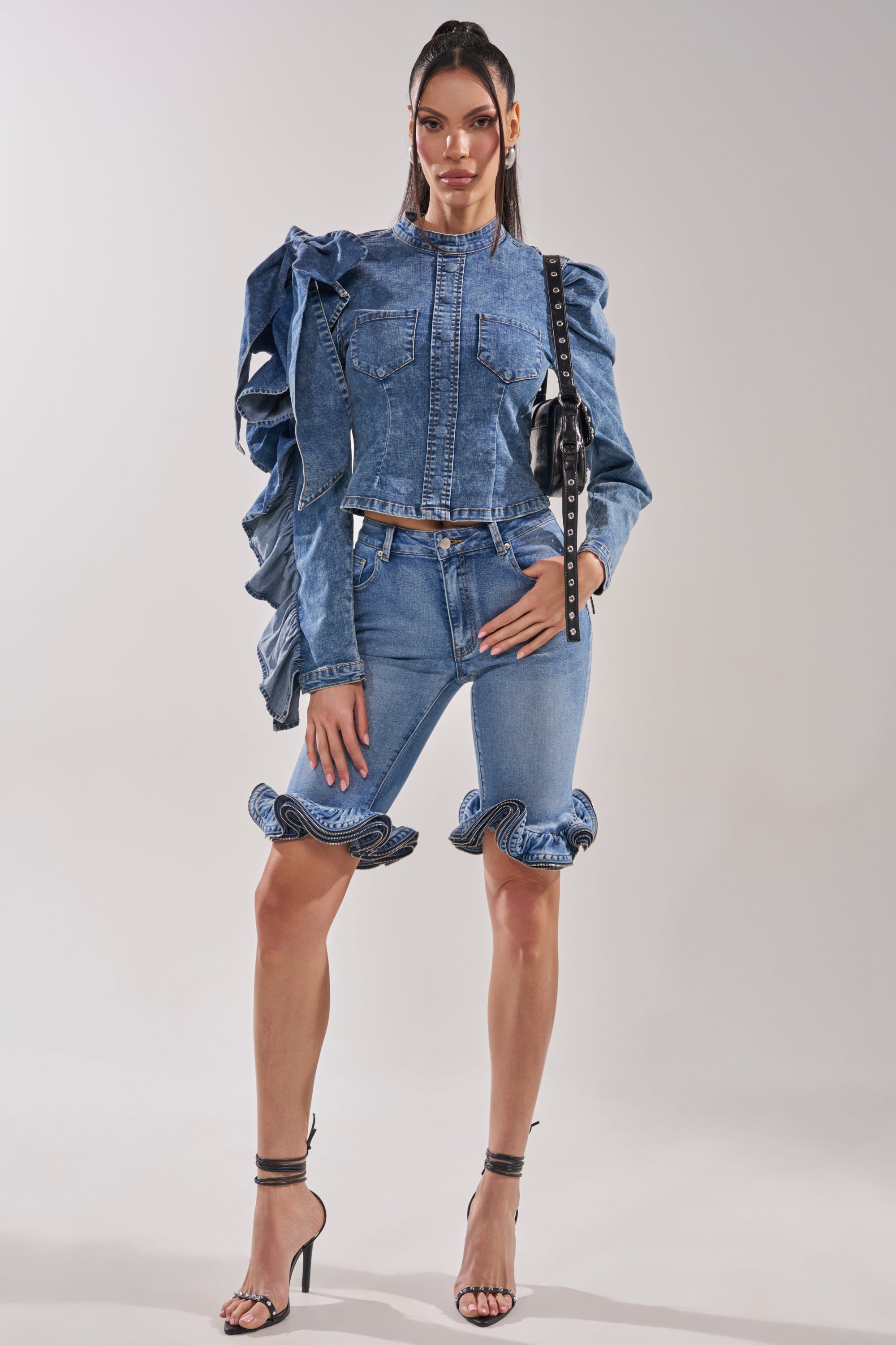 A woman wears the SEIZE THE DAY LONG SLEEVE FLOUNCE MEDIUM BLUE DENIM TOP with ruffled sleeves, matching knee-length shorts with ruffled hems, black strappy heels, a black bag, and has long straight hair.