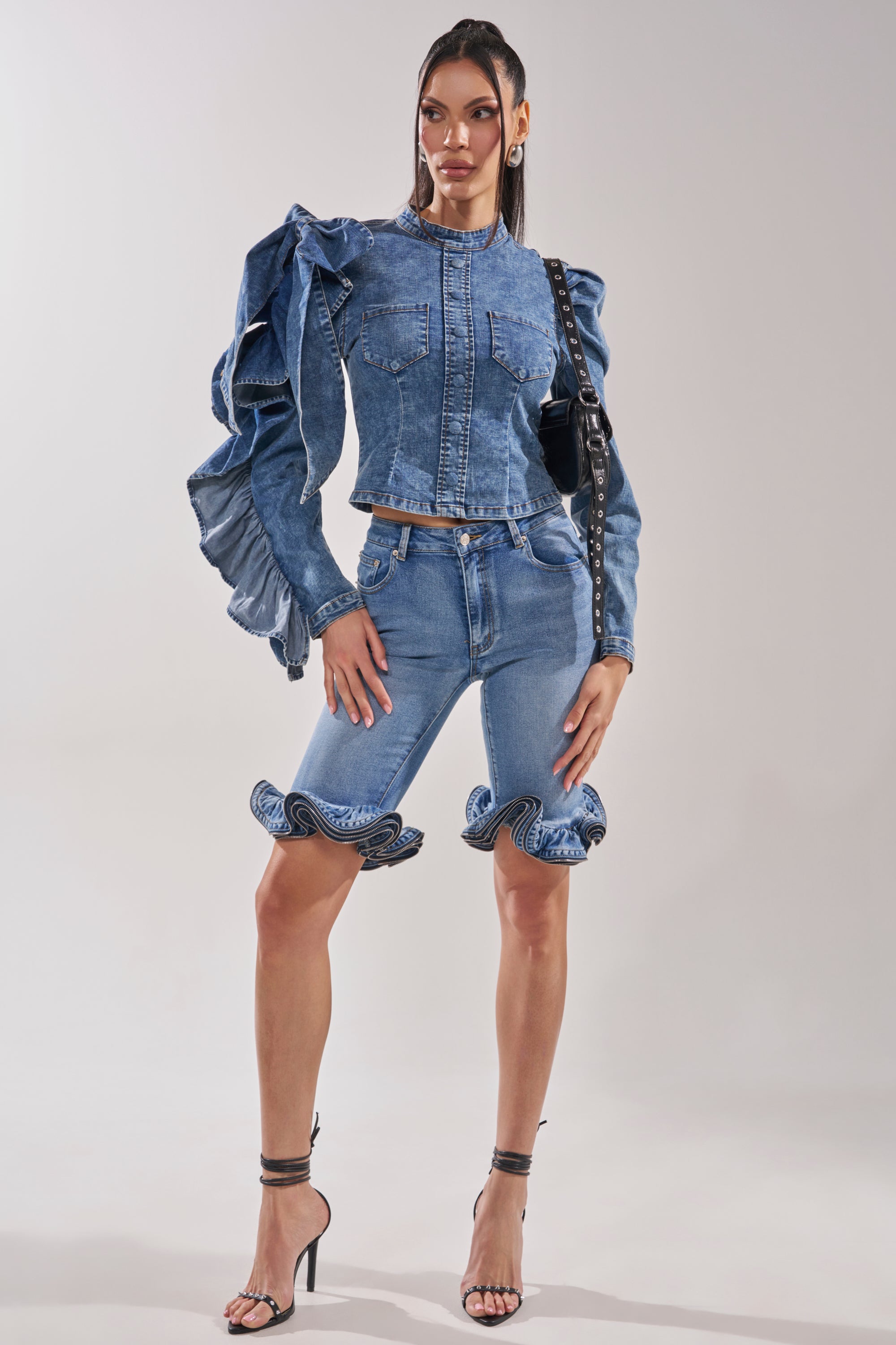 A woman poses in the SEIZE THE DAY LONG SLEEVE FLOUNCE MEDIUM BLUE DENIM TOP, featuring dramatic ruffled sleeves, styled with knee-length ruffled-hem shorts, black high-heeled sandals, and a black shoulder bag against a plain background.