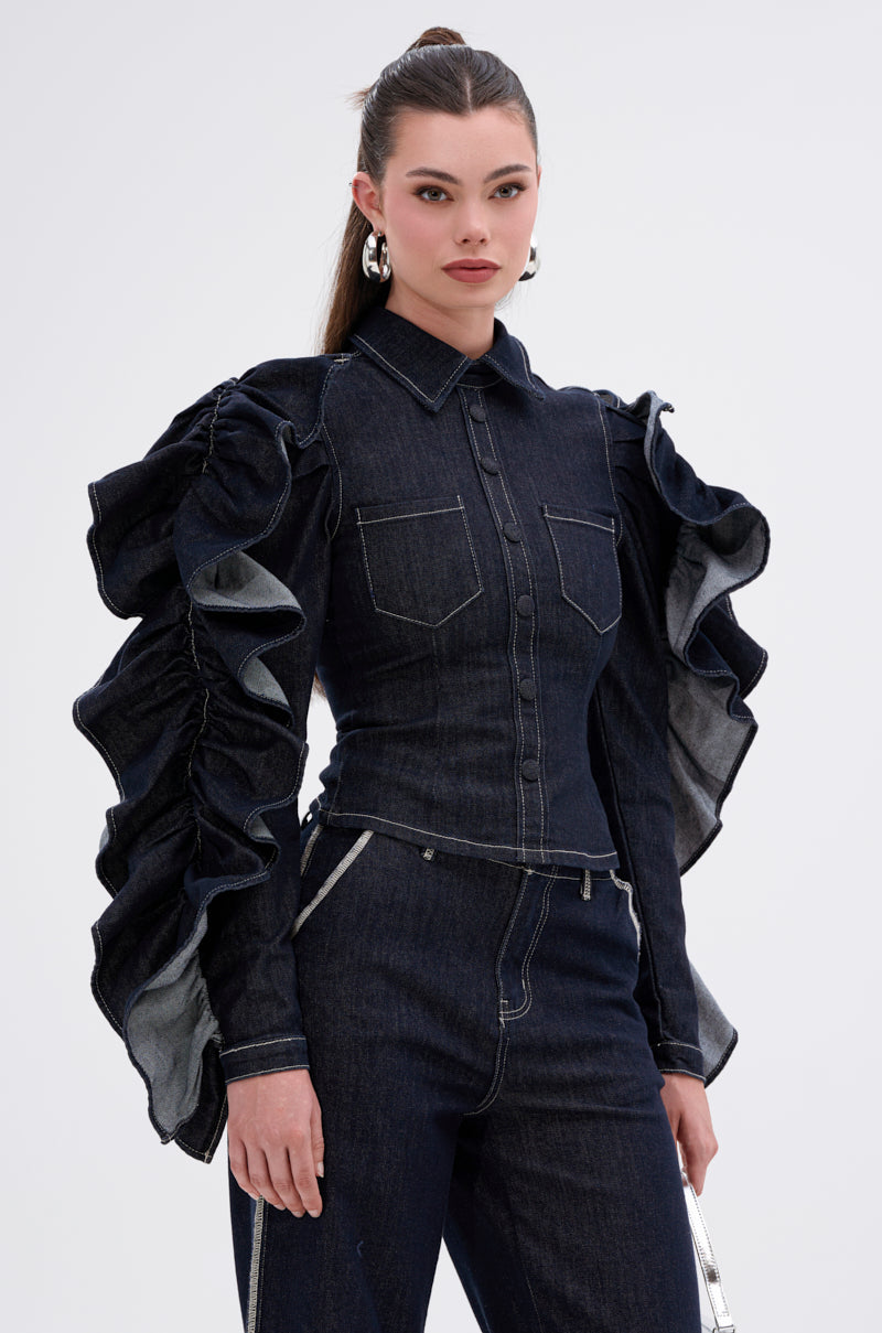 A woman wears the RUFFLED UP LONG SLEEVE DENIM TOP with dramatic ruffle sleeves and chest pockets, paired with matching dark-wash pants featuring zipper details. She accessorizes with silver hoop earrings and has her hair pulled back.