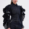 A woman wears the RUFFLED UP LONG SLEEVE DENIM TOP with dramatic ruffle sleeves and chest pockets, paired with matching dark-wash pants featuring zipper details. She accessorizes with silver hoop earrings and has her hair pulled back.
