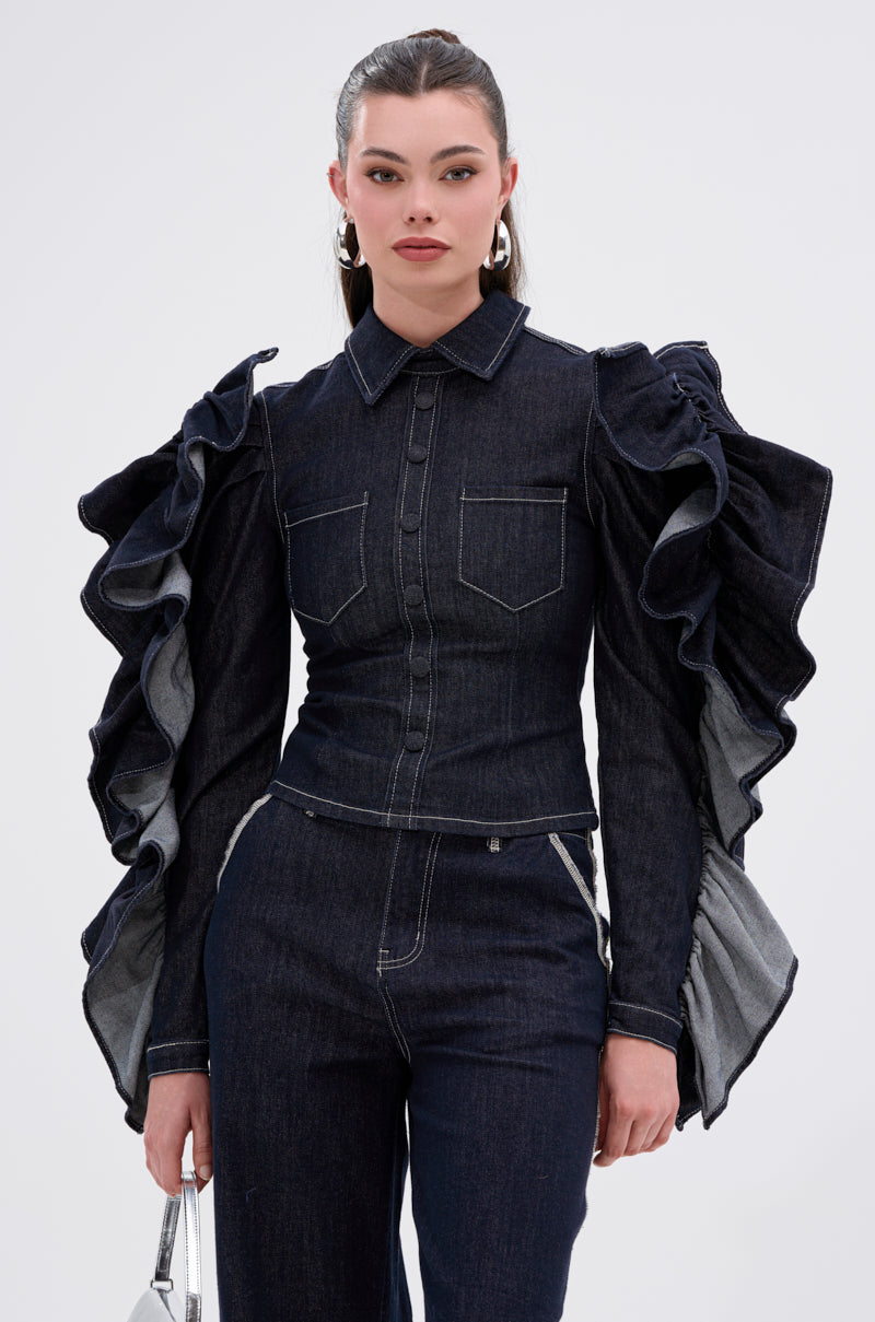 A woman wears the RUFFLED UP LONG SLEEVE DENIM TOP with exaggerated ruffle sleeves and matching pants, holding a small silver handbag. Her hair is pulled back as she stands against a plain light background.