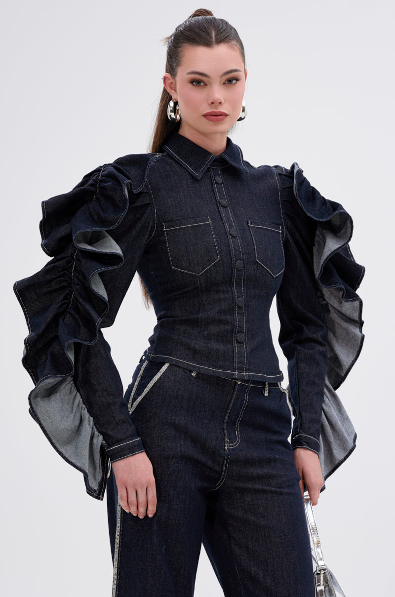 A woman models the RUFFLED UP LONG SLEEVE DENIM TOP, featuring dramatic ruffle sleeves and paired with matching denim pants. She stands against a plain light background with her long hair pulled back and large hoop earrings.
