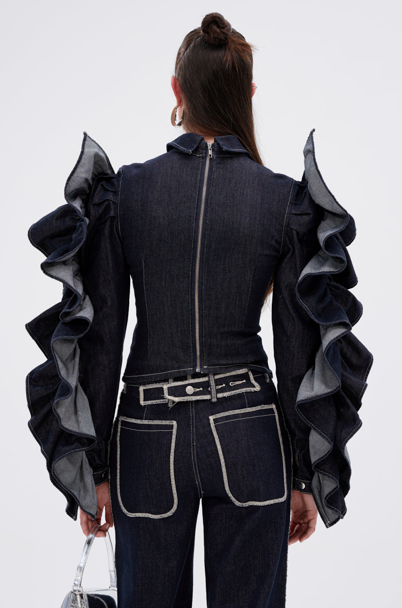 A person stands facing away, wearing the RUFFLED UP LONG SLEEVE DENIM TOP—a dark-wash denim piece with dramatic ruffle sleeves, contrast stitching, a back zipper, belted waist, and holding a small silver handbag.
