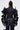 A person stands facing away, wearing the RUFFLED UP LONG SLEEVE DENIM TOP—a dark-wash denim piece with dramatic ruffle sleeves, contrast stitching, a back zipper, belted waist, and holding a small silver handbag.