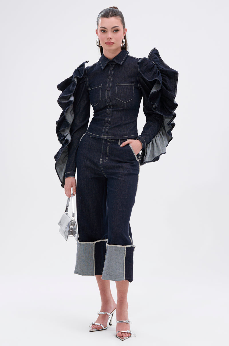 A woman stands against a white background in the RUFFLED UP LONG SLEEVE DENIM TOP with dramatic ruffle sleeves, paired with cropped wide-leg jeans, a small silver purse, and matching silver high-heeled sandals.