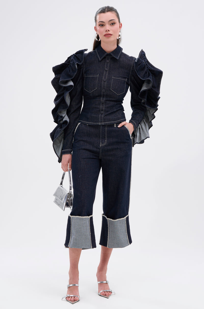 A woman wears the RUFFLED UP LONG SLEEVE DENIM TOP with dramatic ruffle sleeves, paired with dark-wash denim pants, silver heels, and a small silver handbag, standing against a plain white background.