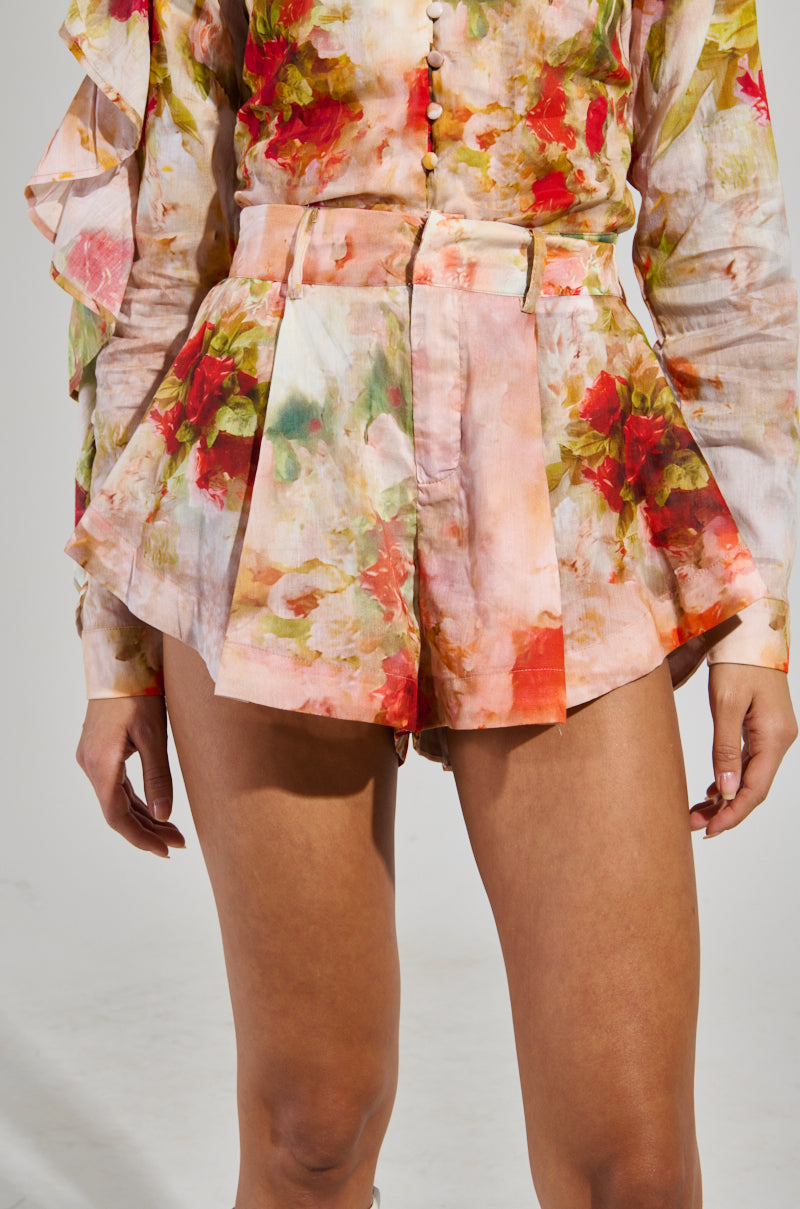 A person wearing a vibrant floral outfit with ruffled sleeves and the Conceited AF Floral Fashion Shorts. This high-rise ensemble features warm patterns in shades of pink, red, and green on a soft background, accentuating the torso and legs.