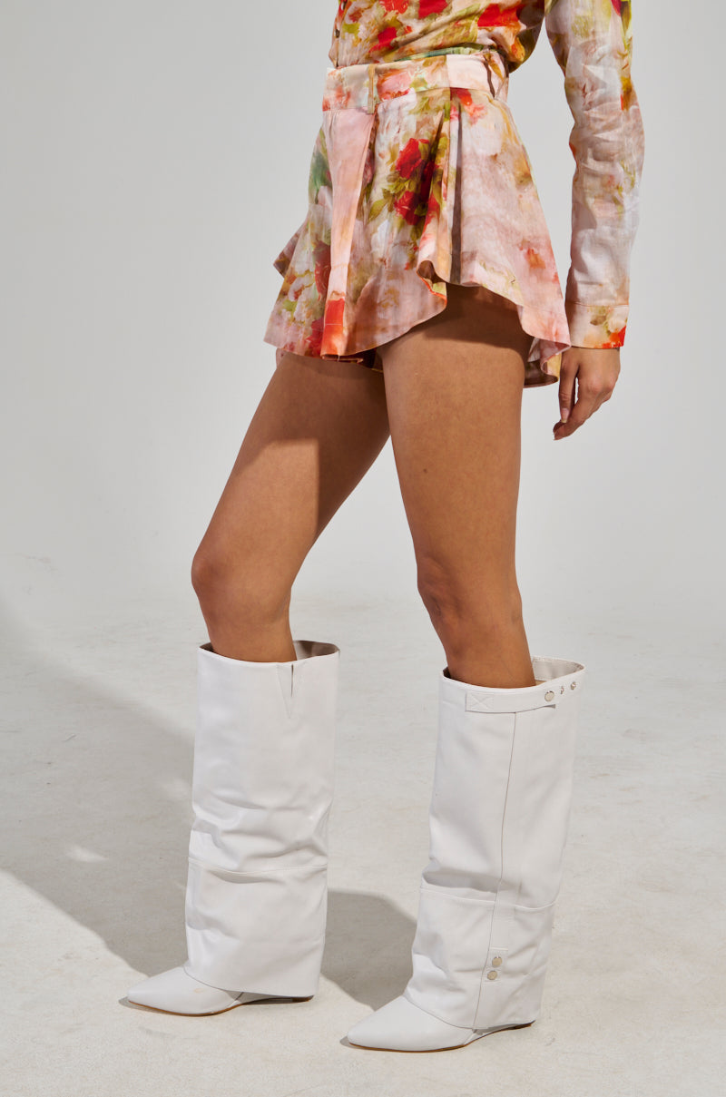 Wearing the stylish Conceited AF Floral Fashion Short ensemble, this look includes a ruffled skirt paired with a coordinating top. Complemented by large, white knee-high slouchy boots with pointed toes, the outfit radiates high-end style against a simple and well-lit backdrop.