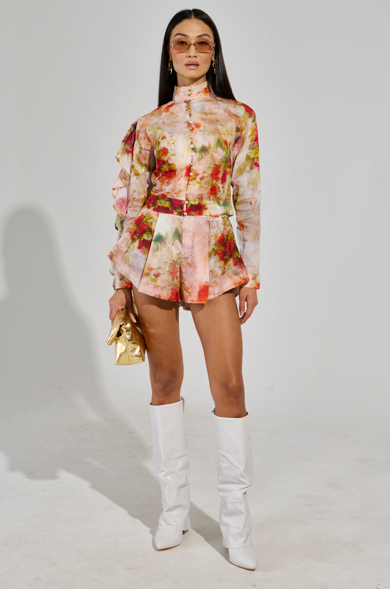 Wearing the CONCEITED AF FLORAL FASHION SHORT romper with ruffled sleeves, paired with white knee-high boots, a person emanates high-rise style. They confidently pose against a plain background while holding a gold clutch and wearing sunglasses.