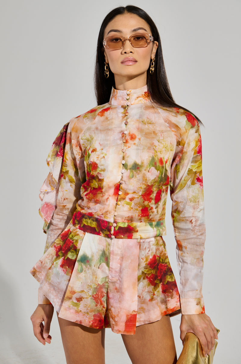 A person wearing the CONCEITED AF FLORAL BLOUSE featuring ruffled sleeves, along with matching high-waisted shorts in a cropped silhouette, poses confidently. They have long hair, sunglasses, and gold earrings while holding a metallic clutch against a plain light gray background.