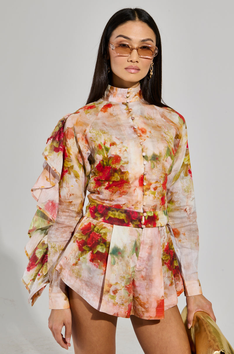 A person wearing the CONCEITED AF FLORAL BLOUSE, which features vibrant floral patterns and ruffled sleeves, poses against a plain background. They pair it with a pleated skirt, have long dark hair, and are sporting sunglasses, effortlessly flaunting their cropped silhouette.