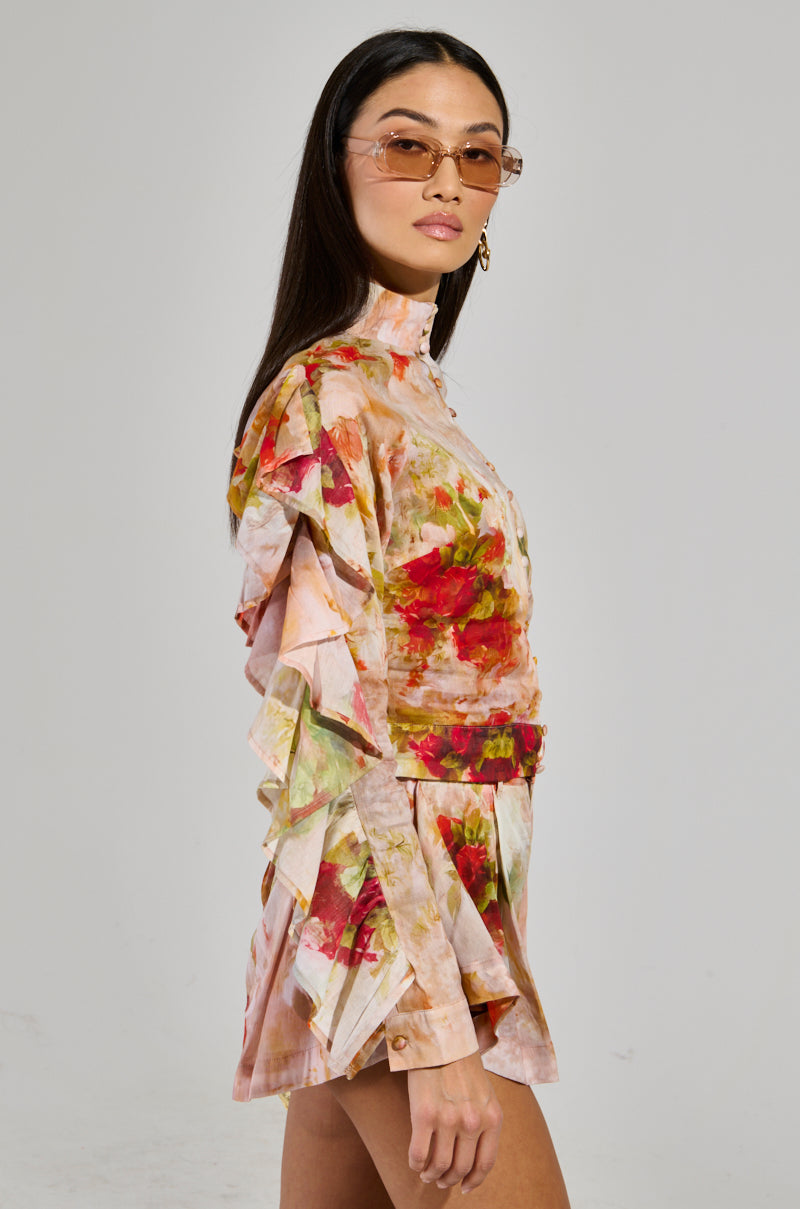 Dressed in the effortlessly stylish CONCEITED AF FLORAL BLOUSE, a person wearing pink-tinted sunglasses and shorts crafted from light linen stands against a plain background. With long dark hair and a neutral expression, they showcase the blouse's ruffled sleeves and cropped silhouette elegantly.