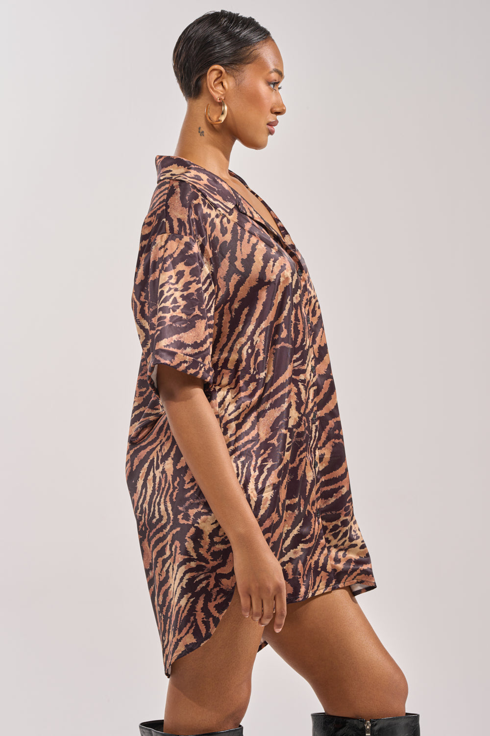 A woman in profile wears the DON'T MAKE A SOUND PRINTED MINI DRESS IN TIGER—a loose, short-sleeved mini. She has short slicked-back hair, gold hoops, and knee-high black boots, posing against a plain light background. 2
