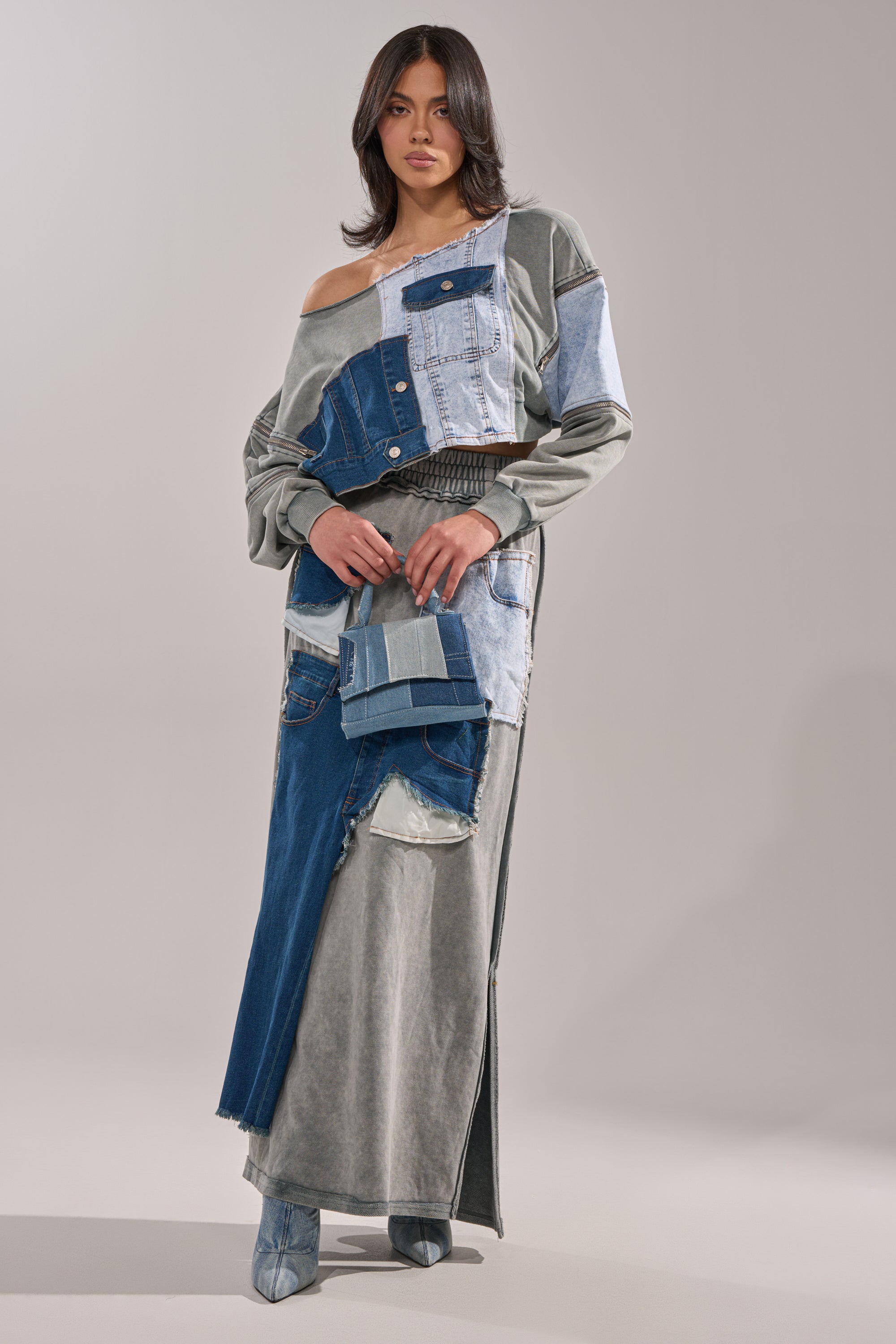 A woman poses against a plain light gray background, wearing the DENIM DREAMS ZIPPER DETAIL MAXI SKIRT with an elastic waistband, a matching crossbody bag, ankle boots, and loose wavy hair.
