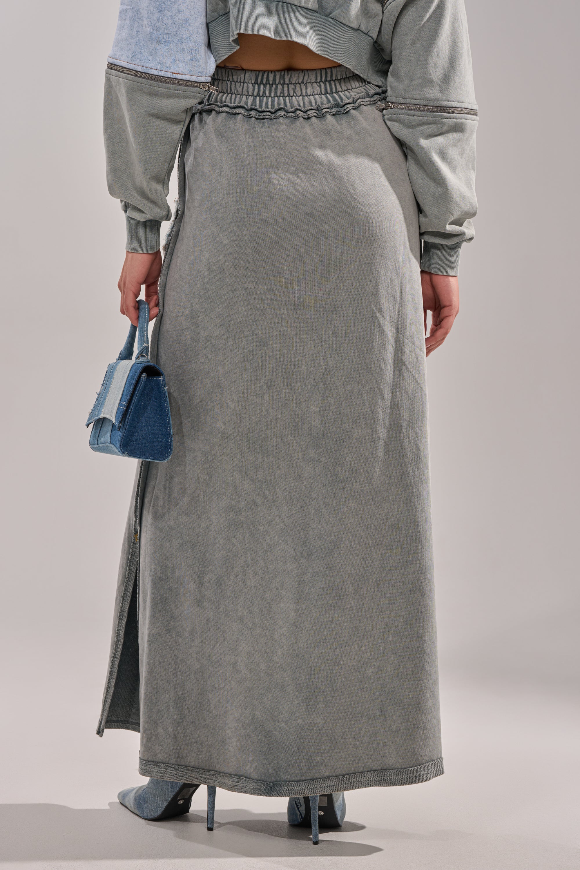 Someone wears the DENIM DREAMS ZIPPER DETAIL MAXI SKIRT, a long high-waisted patchwork denim skirt, with a gray top, blue high-heeled shoes, and holds a small blue handbag. Seen from the back against a plain light background.