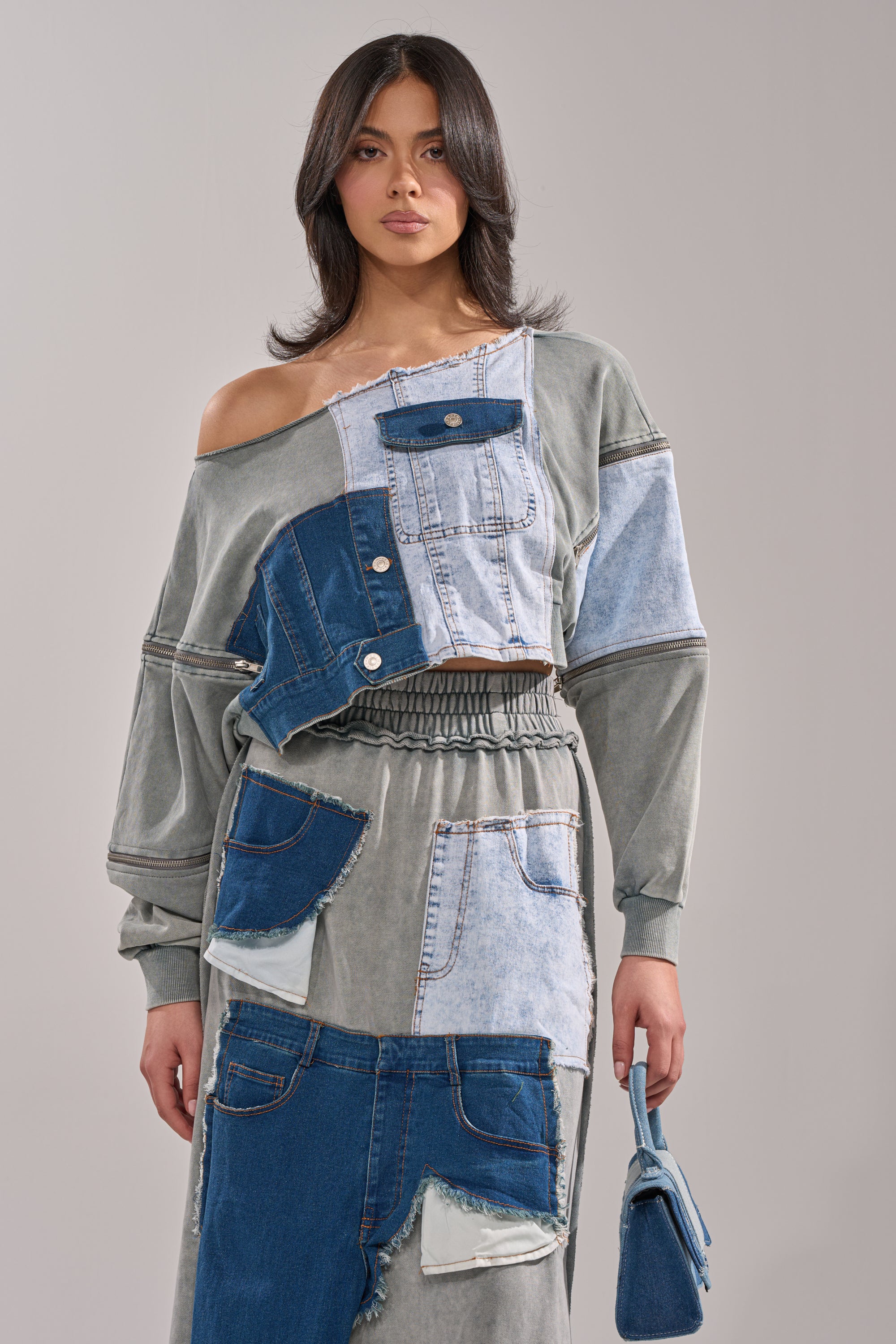 A woman with medium-length dark hair wears the DENIM DREAMS ZIPPER DETAIL SWEATSHIRT in shades of denim and gray, featuring pockets, buttons, and a subtle zipper detail. She finishes the look with a small blue handbag.