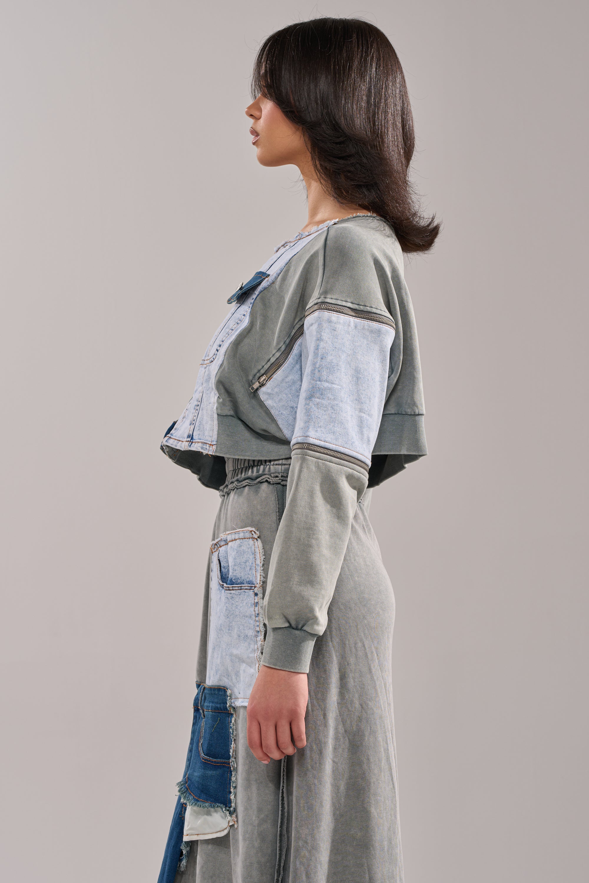 A woman stands in profile wearing the DENIM DREAMS ZIPPER DETAIL SWEATSHIRT, a cropped gray and light blue denim top with patchwork and zipper accents, against a neutral gray background.